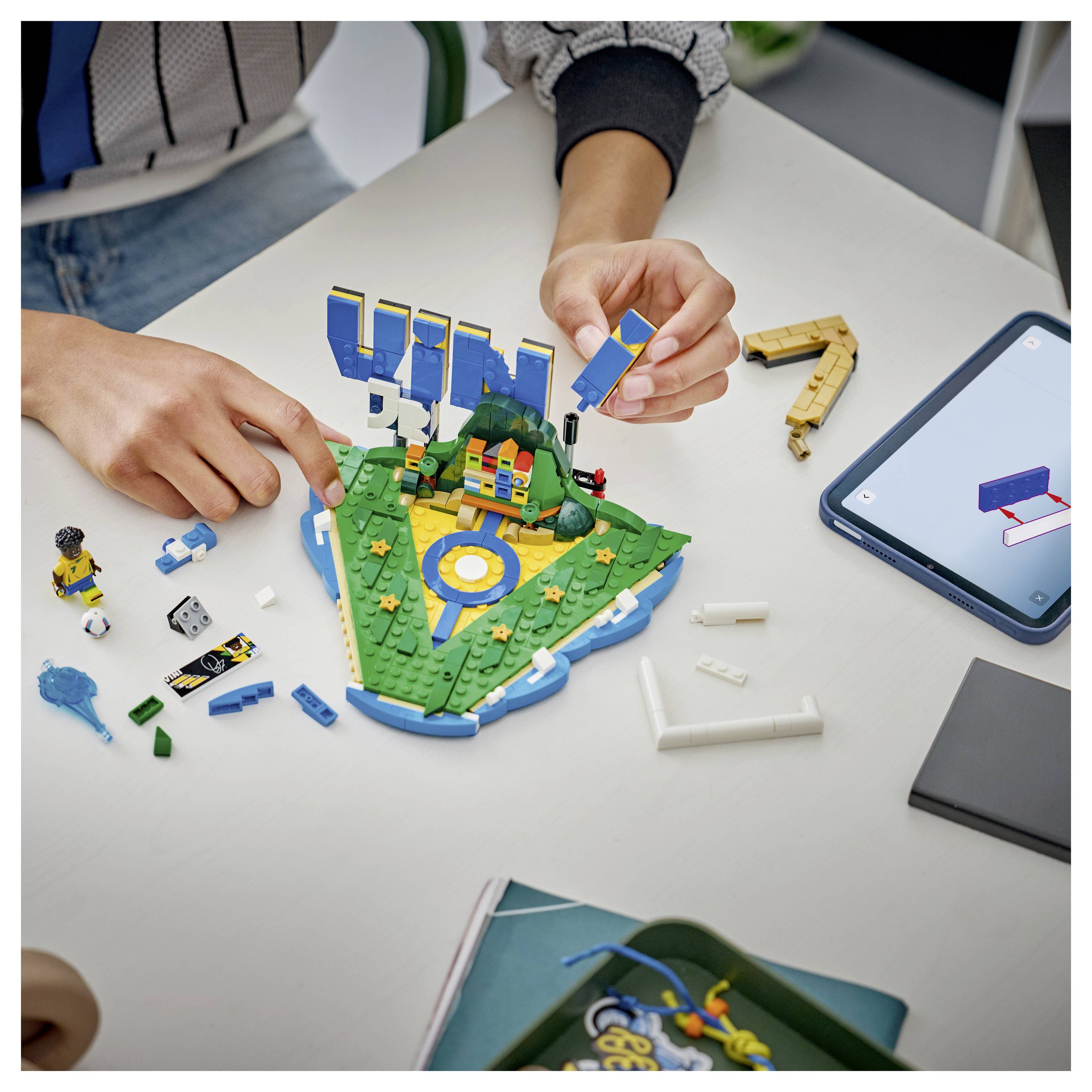Ruce skládající model baseballového hřiště ve stylu LEGO na stole, s volnými kostičkami a tabletem poblíž zobrazujícím návod na stavbu.
