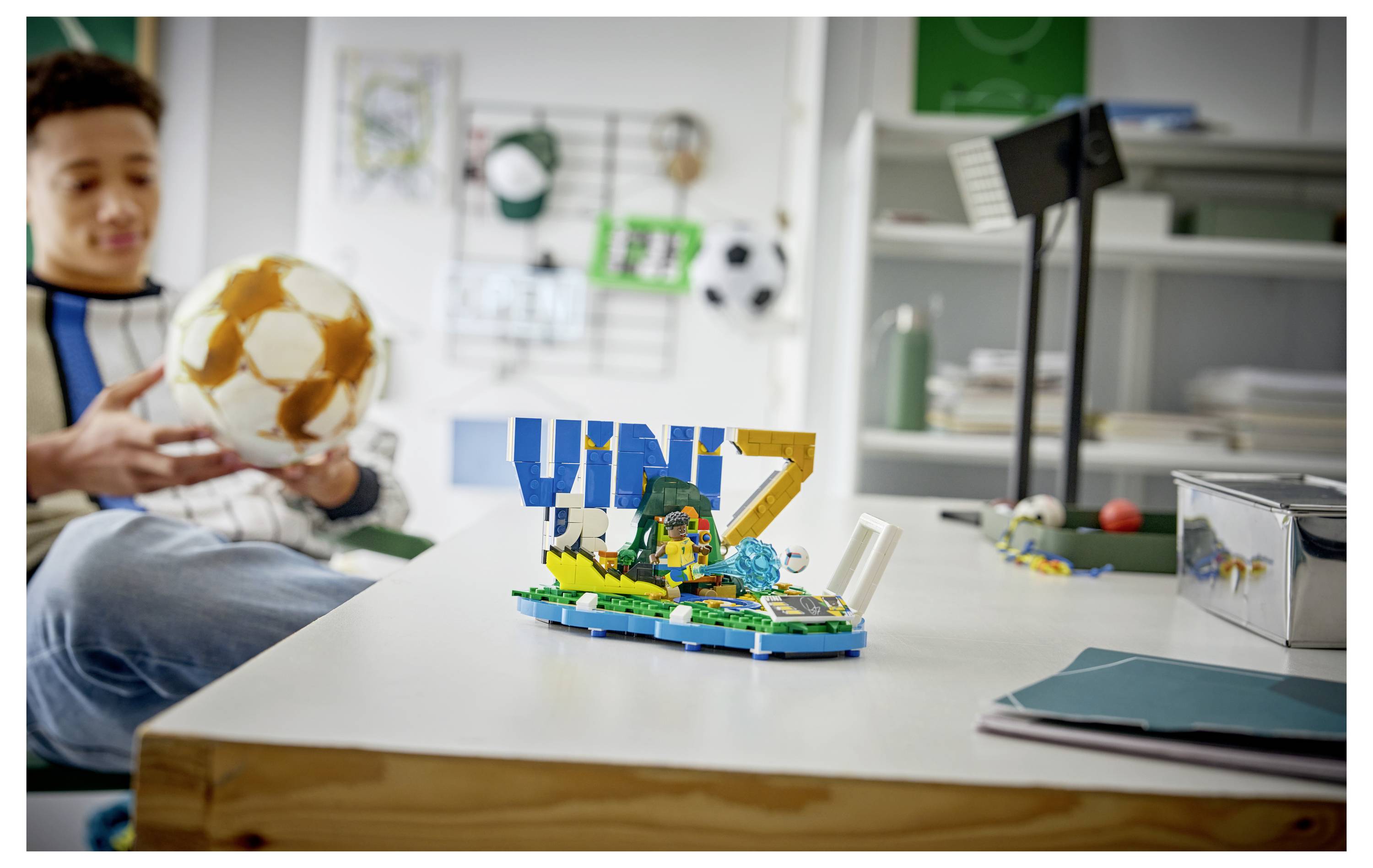 Chlapec v místnosti drží fotbalový míč, zatímco displej ve stylu LEGO na stole ukazuje velká modrá a zlatá písmena s malou scénou.