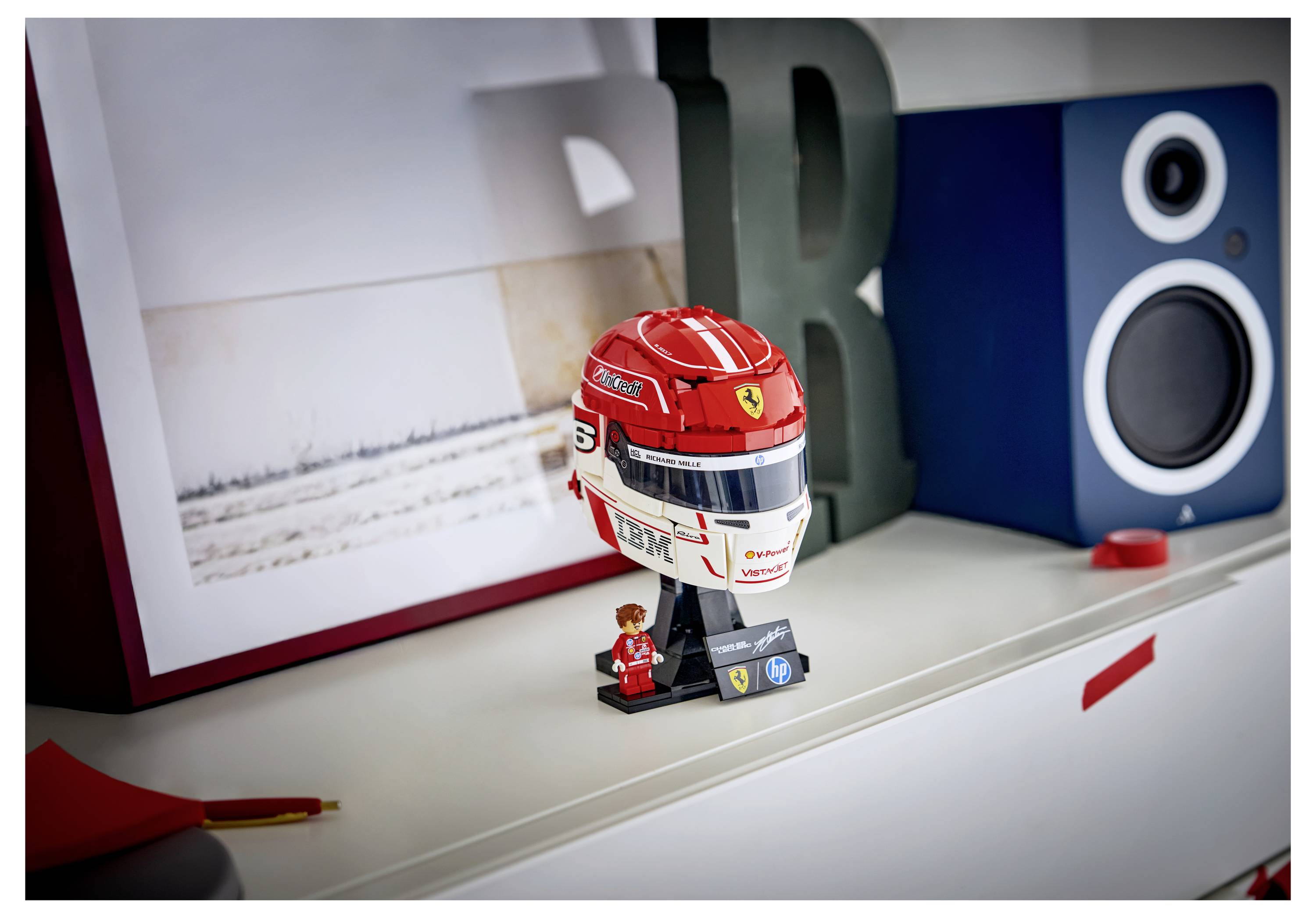 Detailní hračkářská přilba s logem Ferrari a číslem 5 leží na polici poblíž figurky, obklopena fotografickým uměním a reproduktorem.
