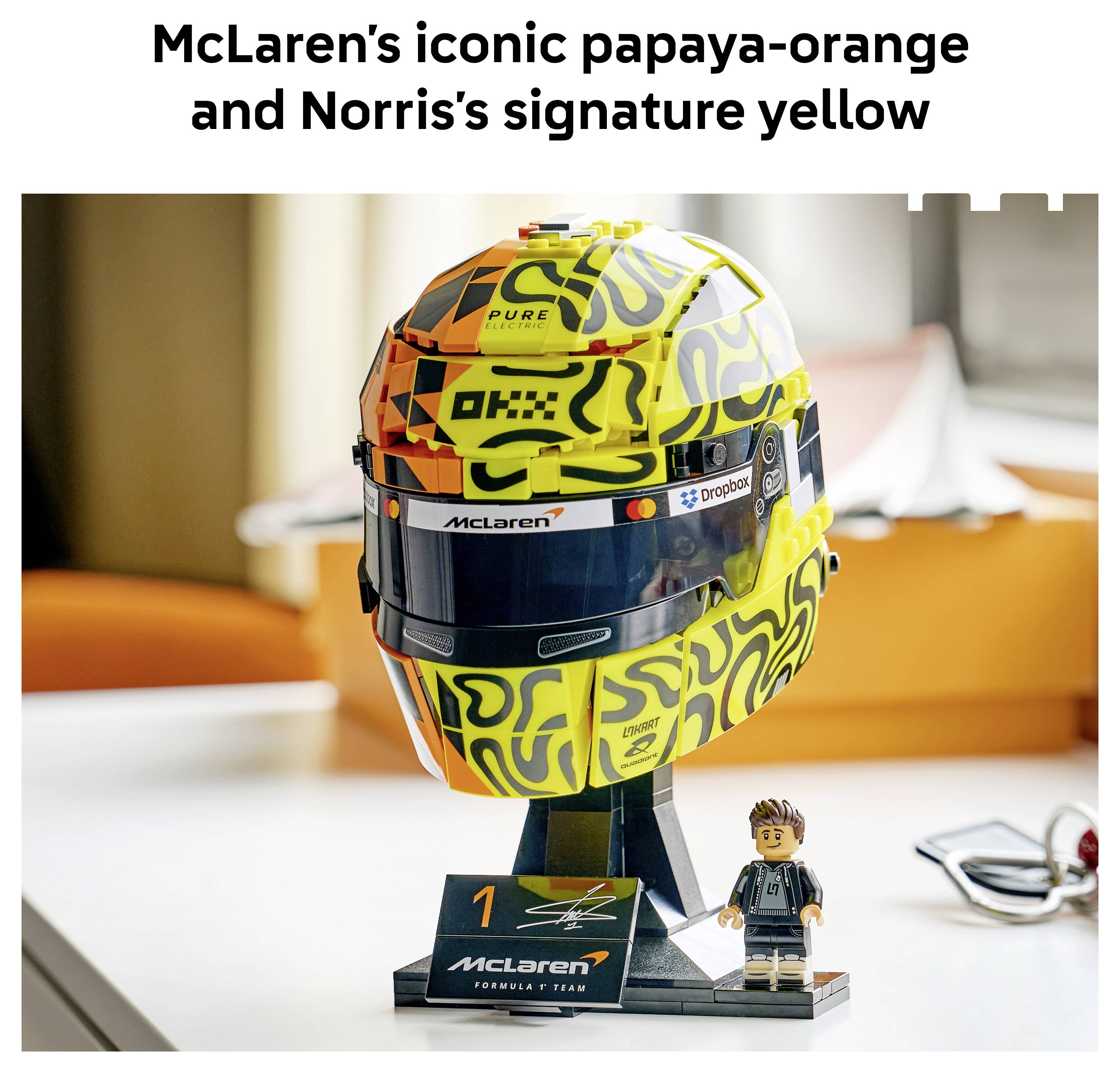 McLarenská helma Formula 1 v oranžovém a žlutém designu vystavená s malou figurkou a týmovou cedulí pod ní.
