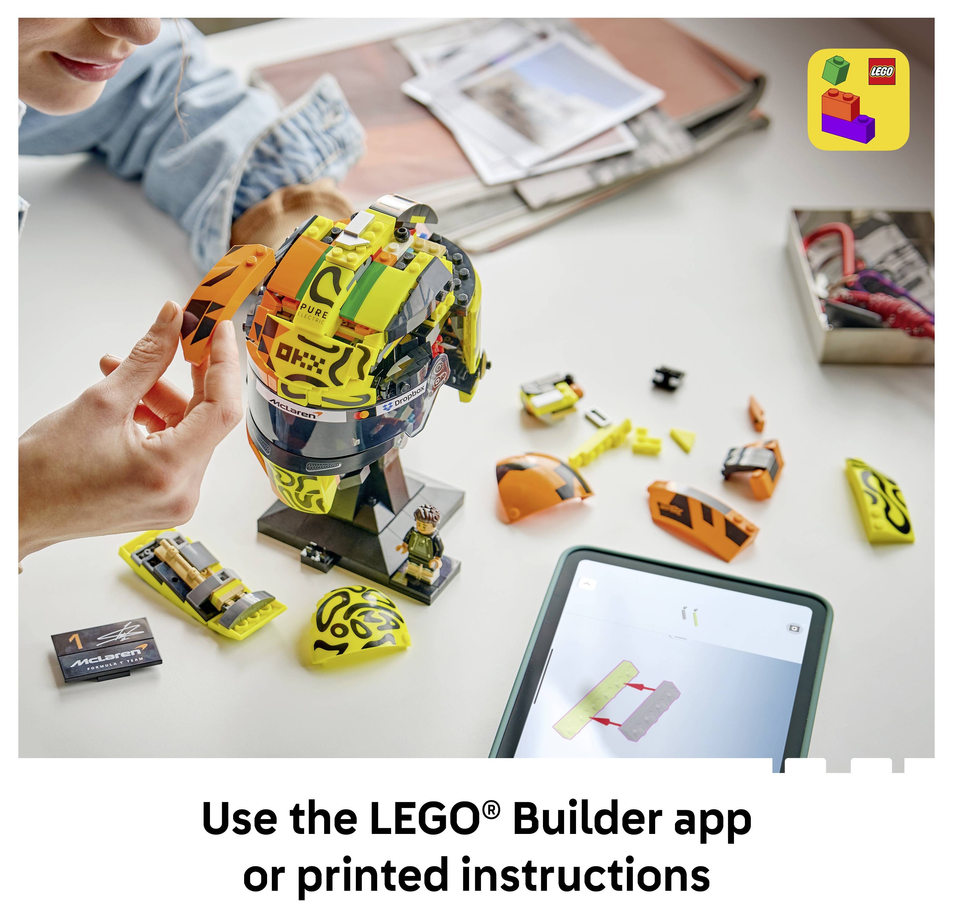 Osoba skládající model závodního helmy LEGO na stole s volnými součástkami a tabletem zobrazujícím pokyny; text říká "Použijte aplikaci LEGO® Builder nebo tištěné pokyny".