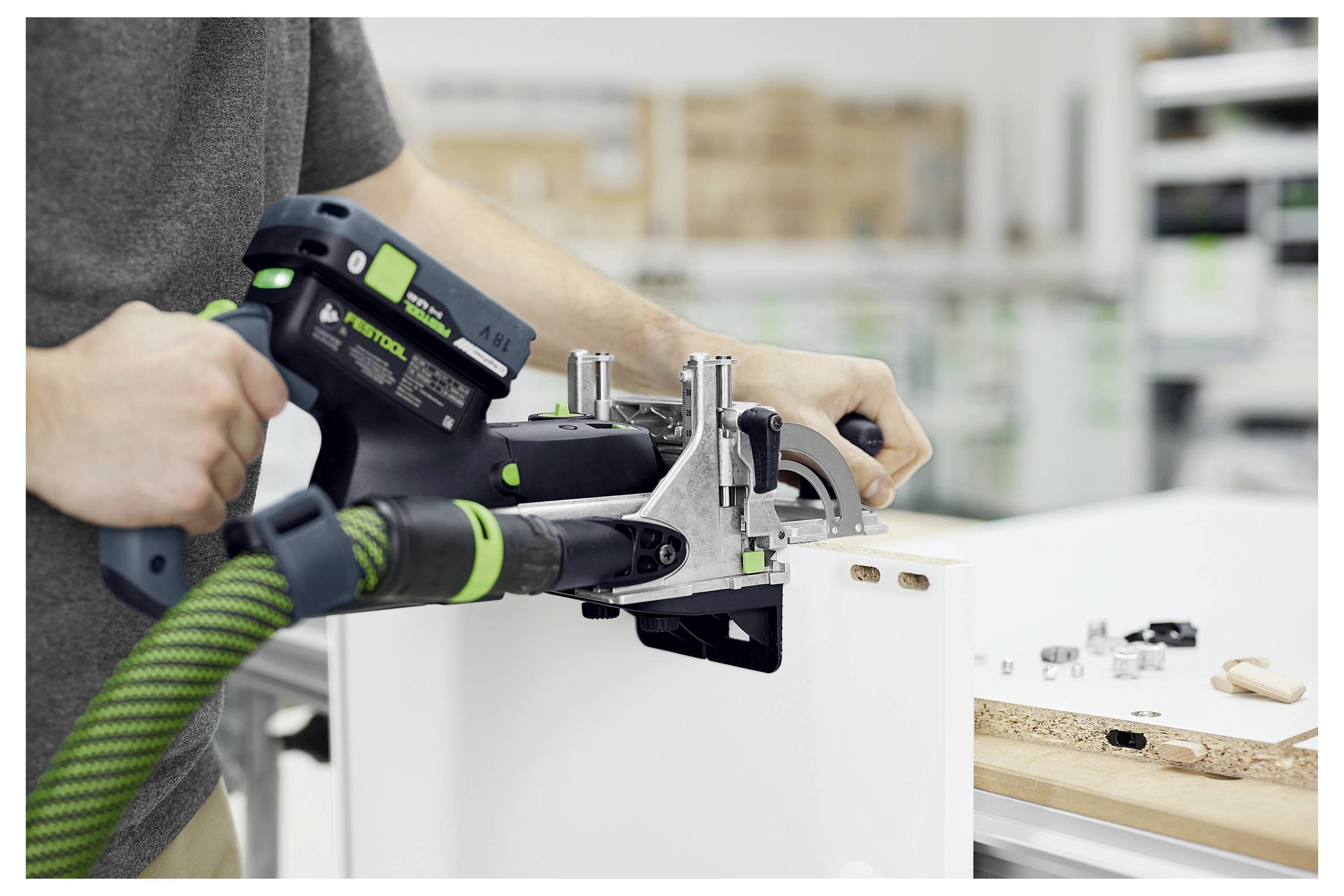 Osoba používající Festool Domino Jointer na dřevěné desce v dílně, obklopená nástroji a kováním, zdůrazňující proces zpracování dřeva.