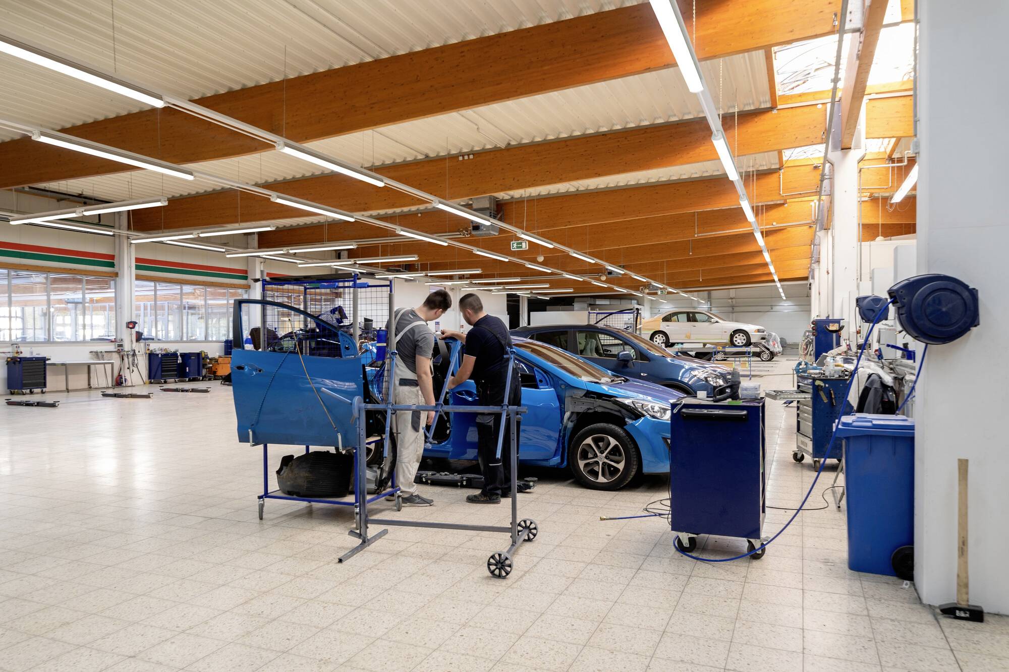 Dva mechanici pracují na autě v prostorné, dobře osvětlené garáži. Kolem nich je rozmístěno množství nástrojů a zařízení, což zdůrazňuje automechanické prostředí.