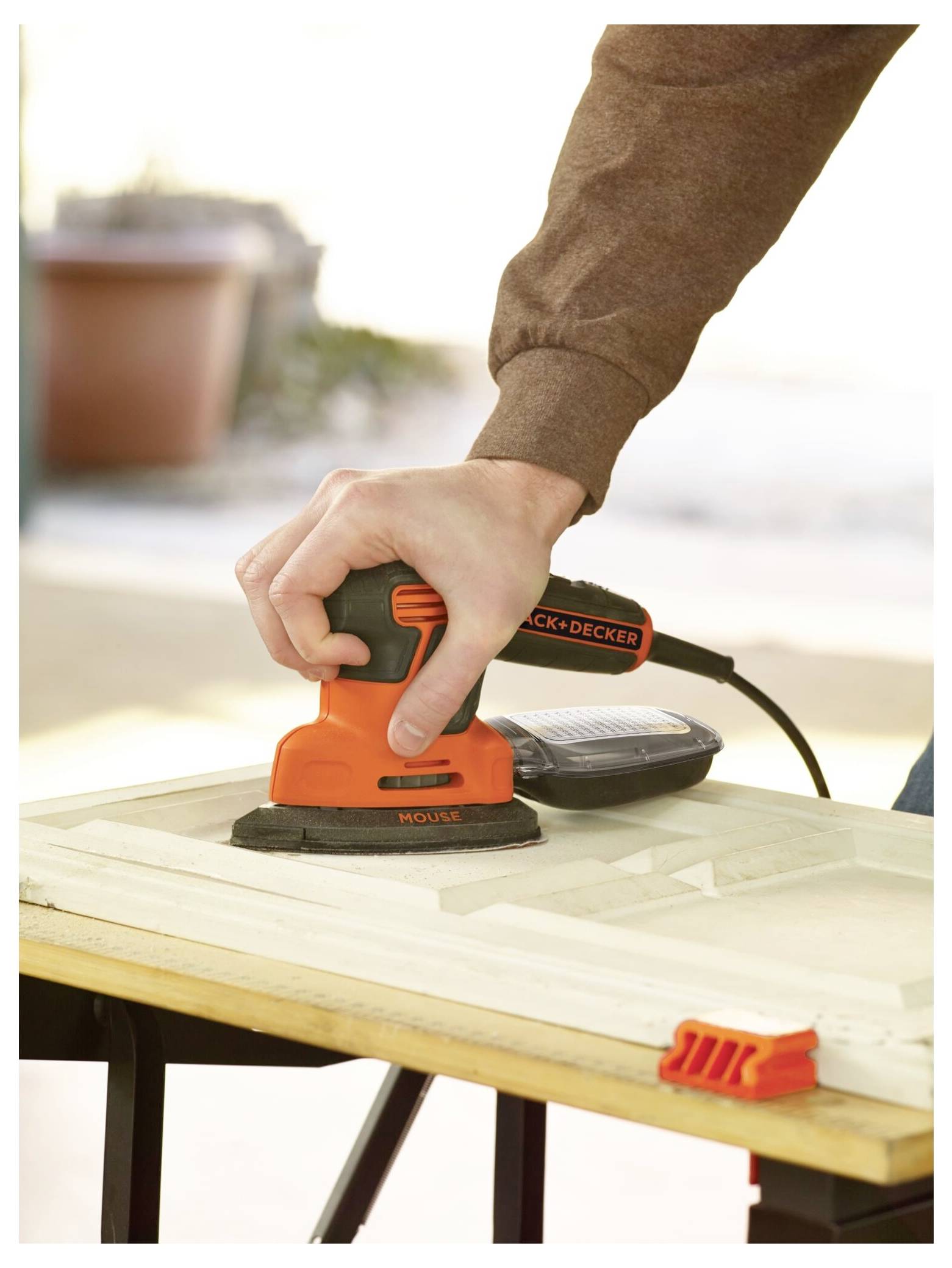 Osoba používá oranžový elektrický vibrátor Black+Decker na dřevěné ploše se zaměřením na úchop ruky a nástroj v provozu.