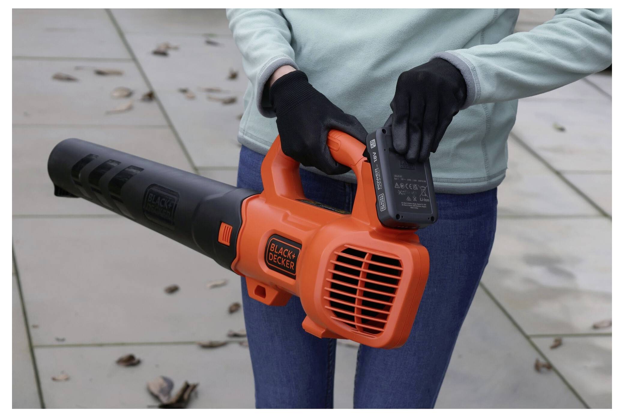 Osoba držící oranžově-černý foukací stroj na listí, která vkládá bateriový balíček, stojící na terase s rozptýlenými listy.
