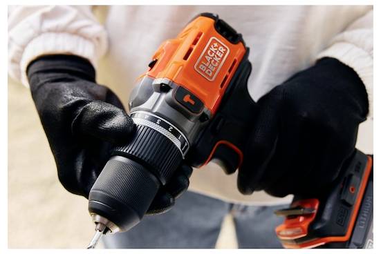 Osoba v černých rukavicích drží oranžovo-černou elektrickou vrtačku značky „BLACK+DECKER", zdůrazňující její úchop a design.