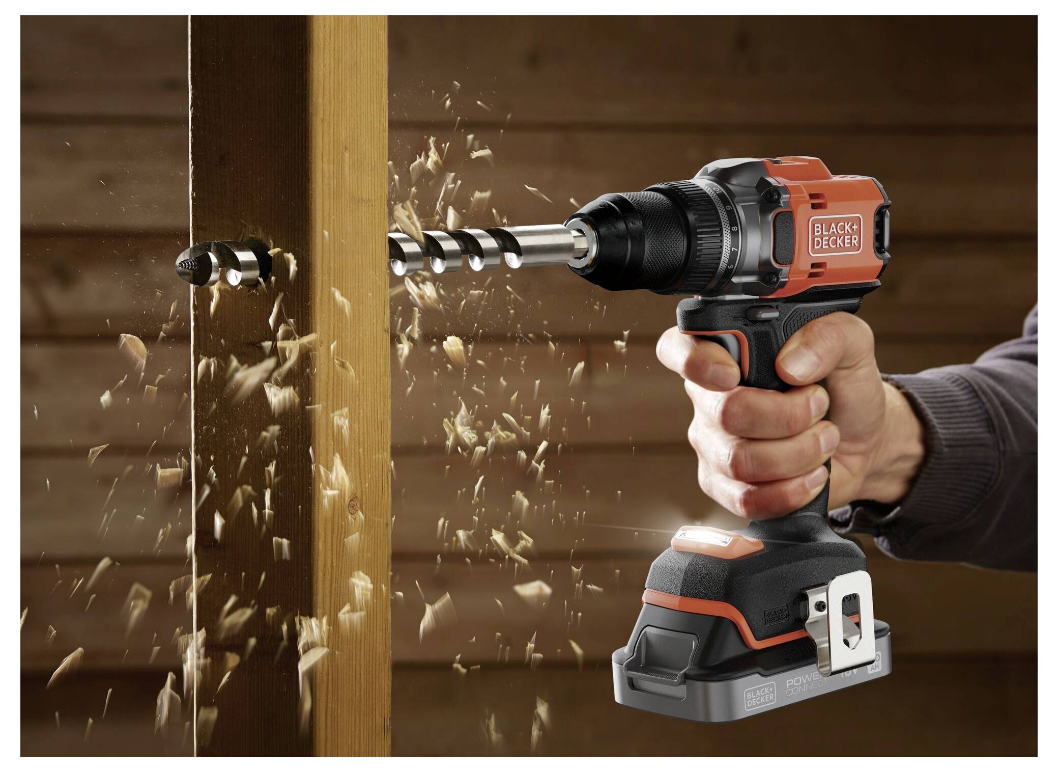 Ruka držící bezdrátovou vrtačku Black & Decker, která vrtá do dřevěné desky, přičemž kolem létají třísky dřeva.