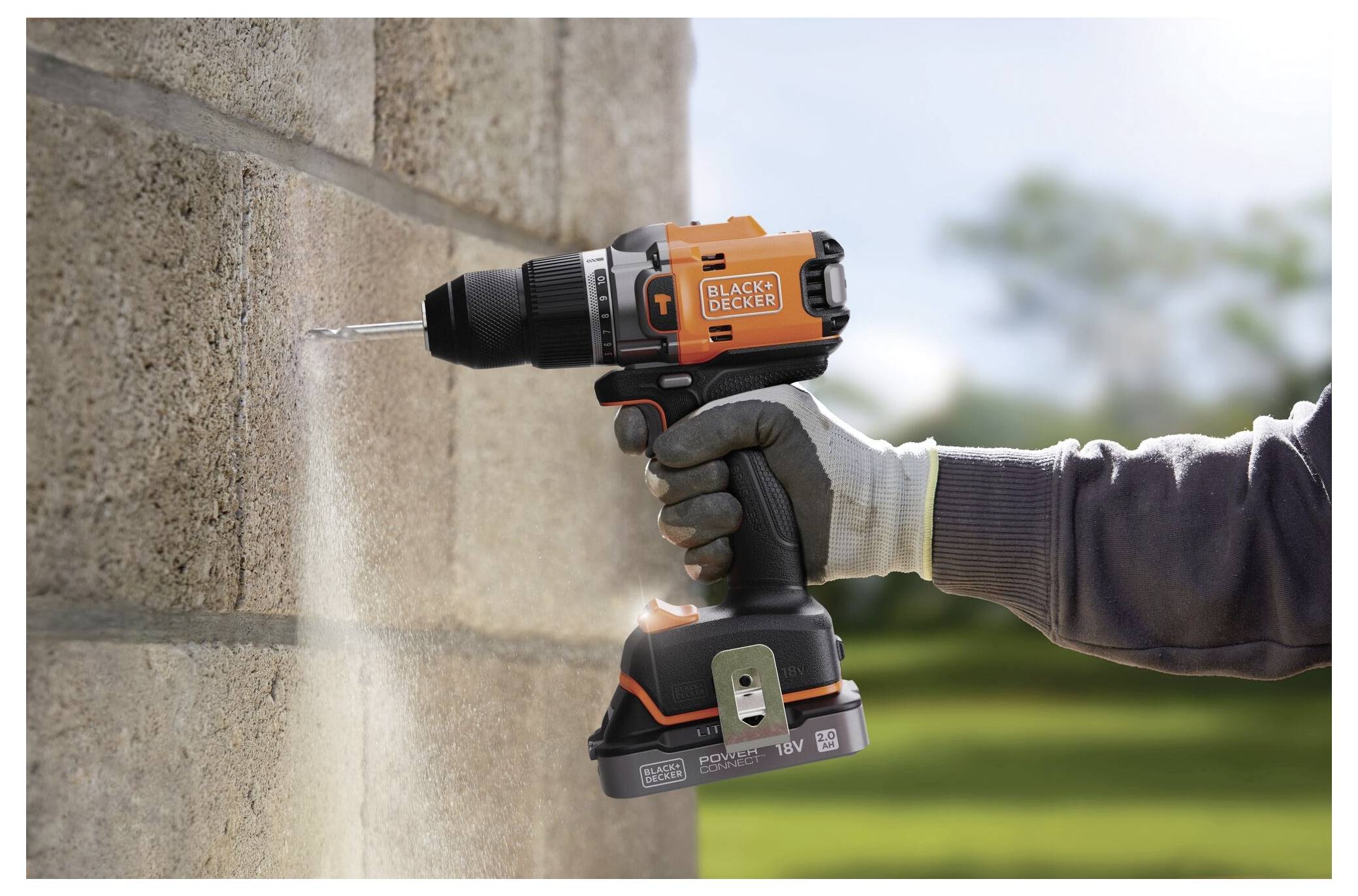Osoba v rukavicích používá vrtačku Black+Decker na vrtání otvoru do cihlové zdi venku. Vrtačka je bezdrátová a oranžová.
