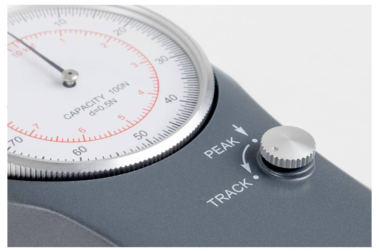 Detailní snímek mechanického číselníkového manometru s kapacitou 100N. Manometr má ovladač označený "PEAK" a "TRACK".