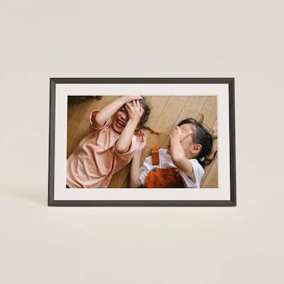 Fotografie v rámu zachycující dva děti ležící na dřevěné podlaze, které se usmívají a hrají si na schovávací s rukama zakrývajícími jejich tváře.