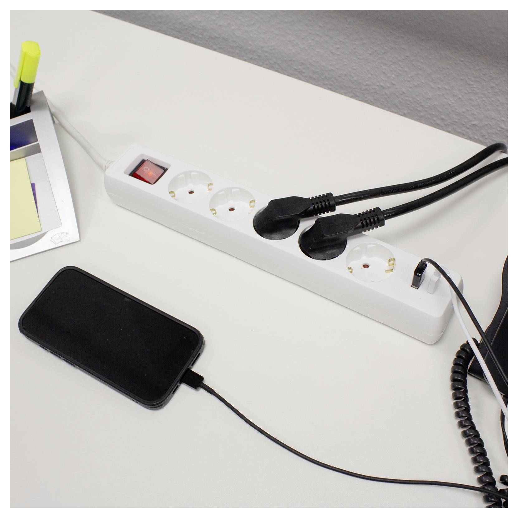 Nabíjení telefonu přes zásuvkový panel na stole s dalšími zapojenými zařízeními, což ukazuje nastavení pracovního prostoru.