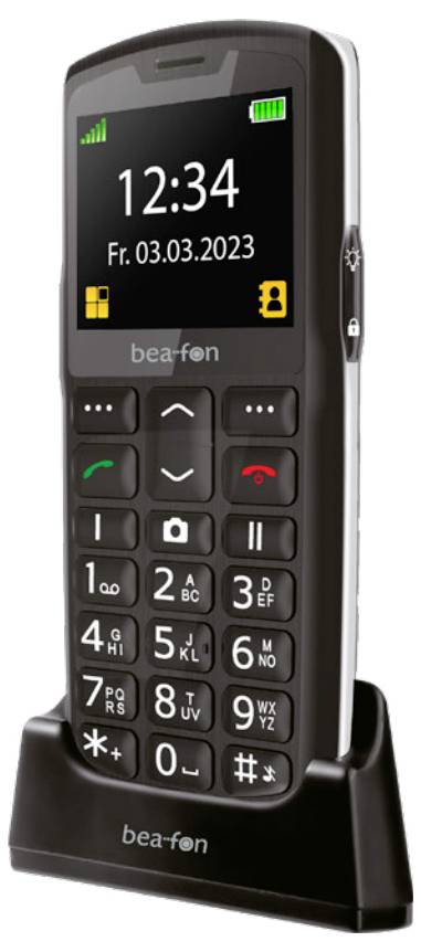 Černý mobilní telefon s velkými tlačítky, malým displejem zobrazujícím „12:34" a datem „Pá 03.03.2023", umístěný na nabíjecím stojanu.