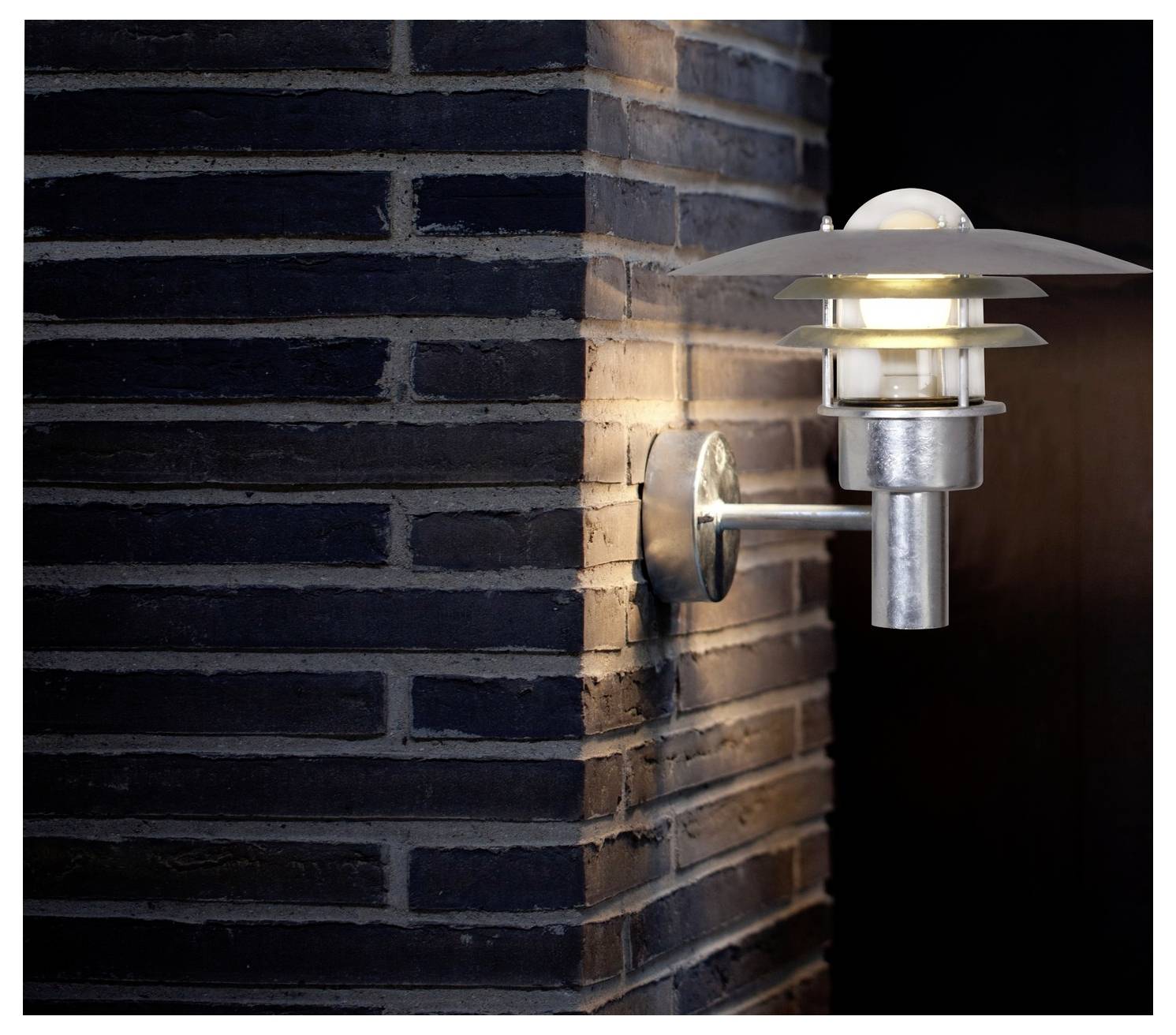 Moderní nástěnná lampa s kovovou úpravou vydává teplé světlo proti tmavé zděné stěně, čímž vytváří útulné a stylové prostředí.