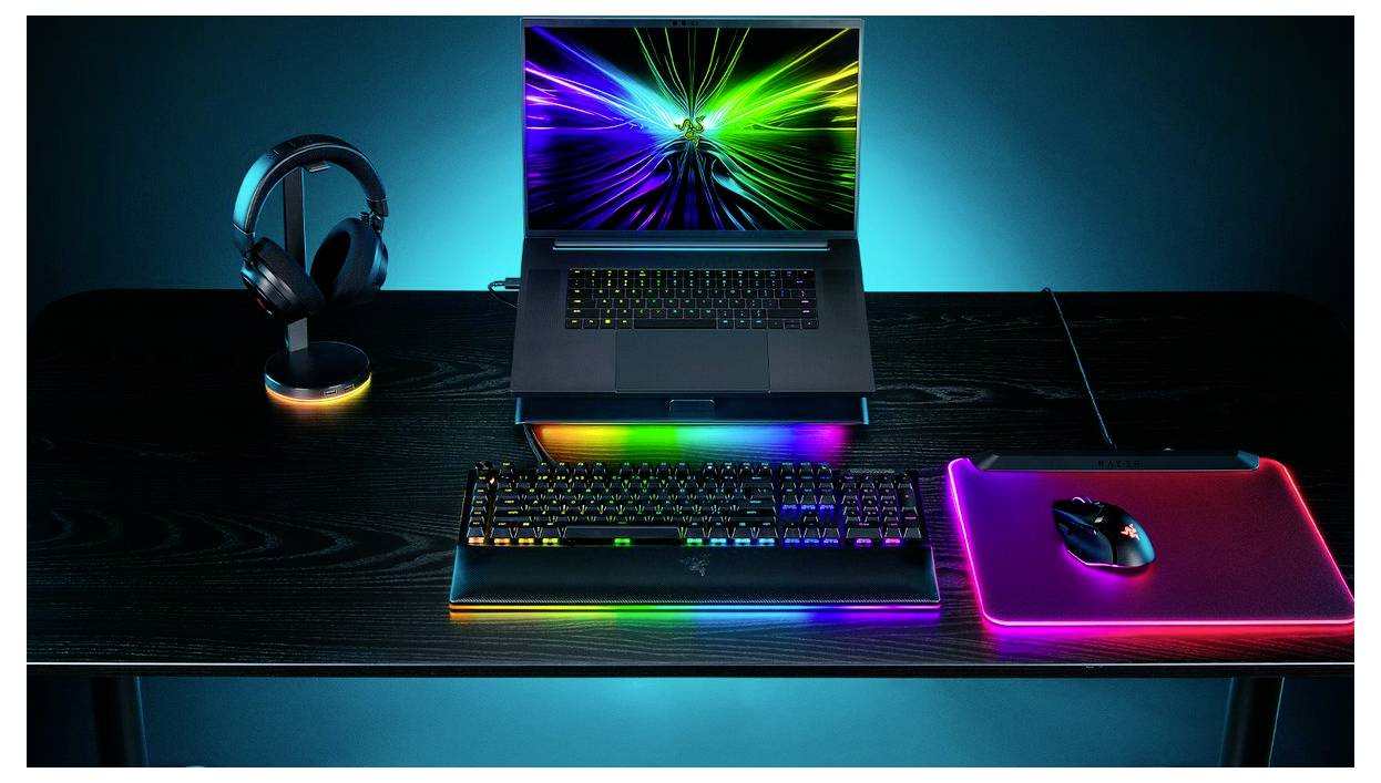 Barevné herní prostředí s notebookem zobrazujícím obrazovku s duhovým vzorem, klávesnicí s RGB osvětlením, myší na osvětleném podložkou a sluchátky.
