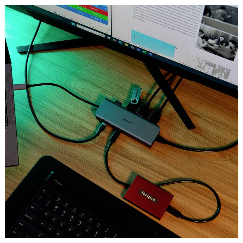 Pracovní prostor se dřevěným stolem obsahuje počítačový monitor, připojenou klávesnici, notebook a různé kabely a adaptéry.