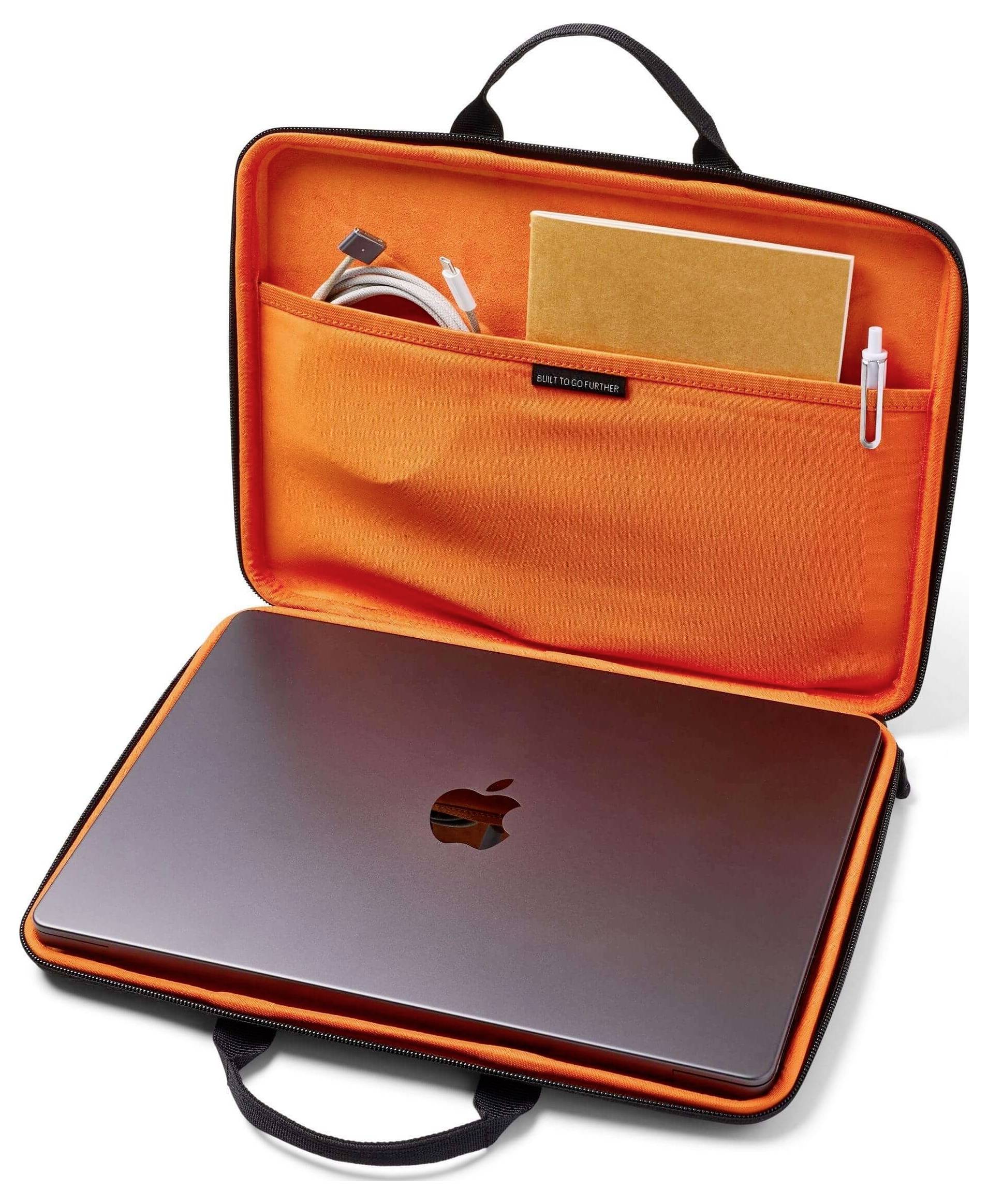 Otevřené pouzdro na notebook s oranžovým interiérem obsahující stříbrný notebook, poznámkový blok, pero a stočený kabel v kapse.