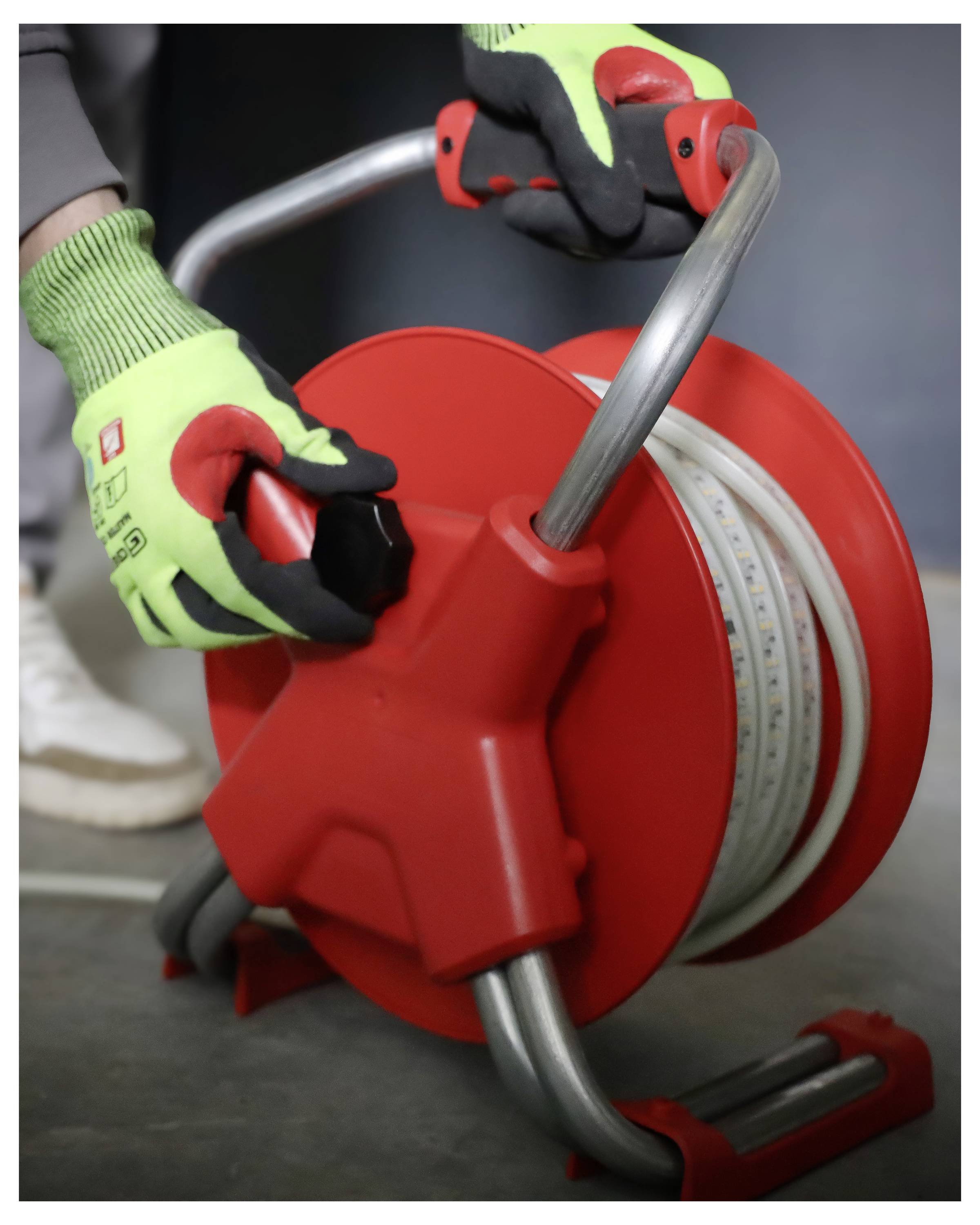 Osoba v zelených rukavicích upravuje červený buben s kabelem a rukojetí, připravený k použití.