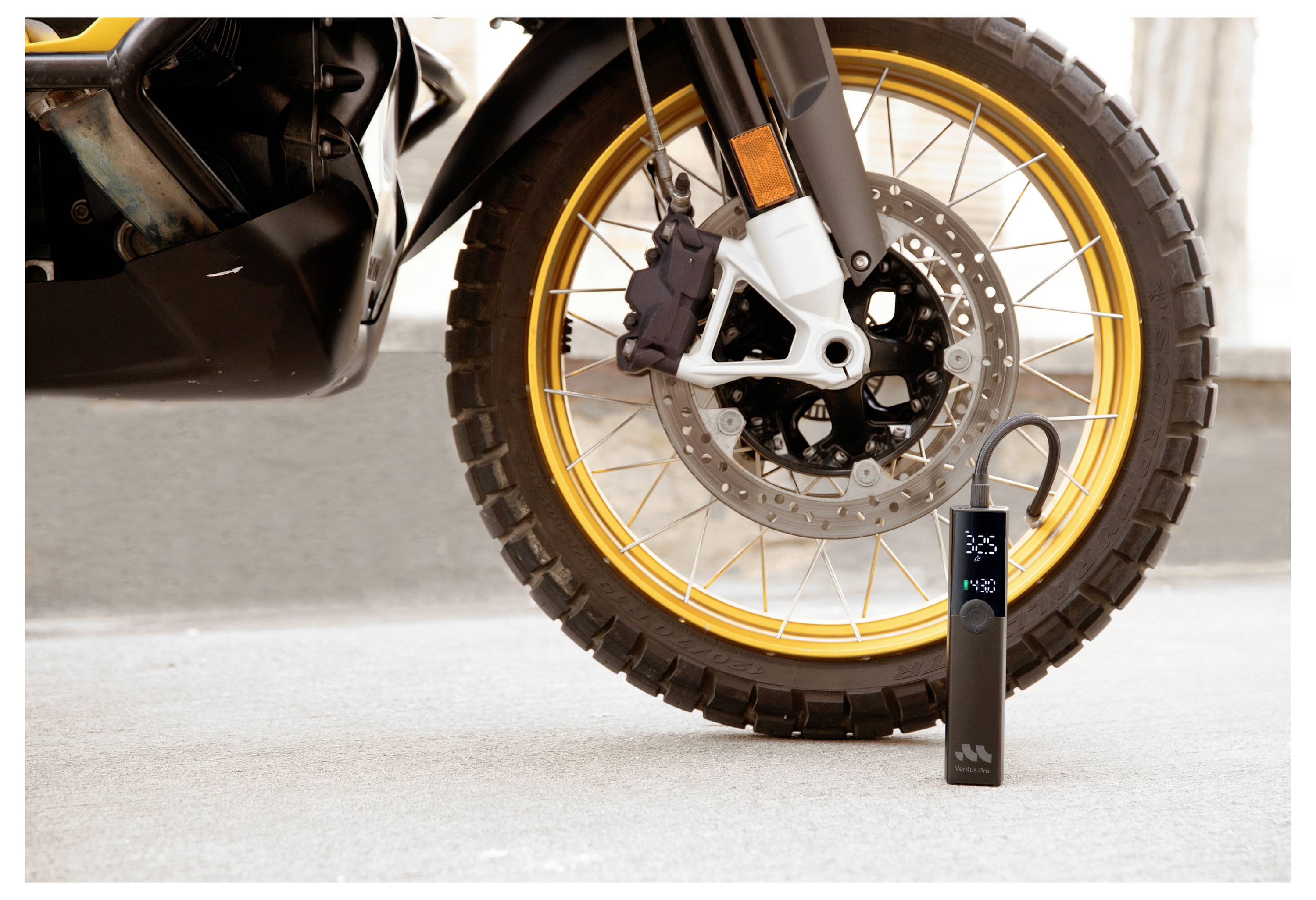 Malý černý digitální manometr na pneumatiky stojí vedle přední kola motocyklu se zlatým ráfkem a ojetým pneu.