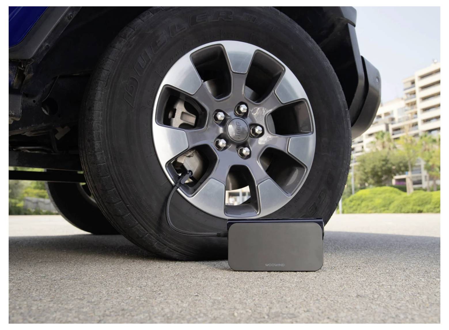 Pneumatika automobilu je téměř prázdná, spočívá na malém přenosném zařízení před kolem, v pozadí se nacházejí budovy a stromy.
