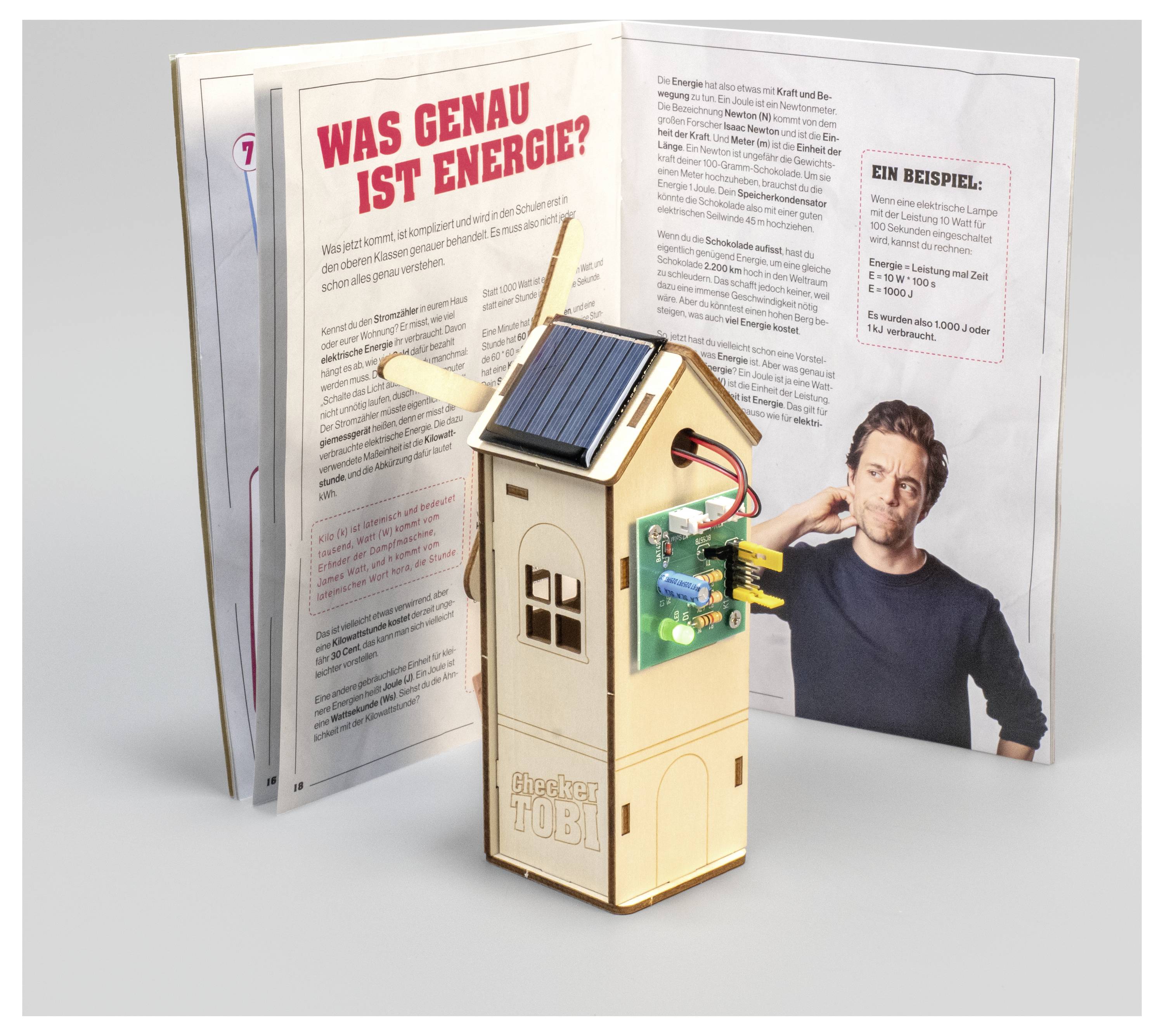 Dřevěný model se solárním panelem a deskou plošných spojů stojí před otevřenou knihou s názvem „Was genau ist Energie?" a fotografií osoby.