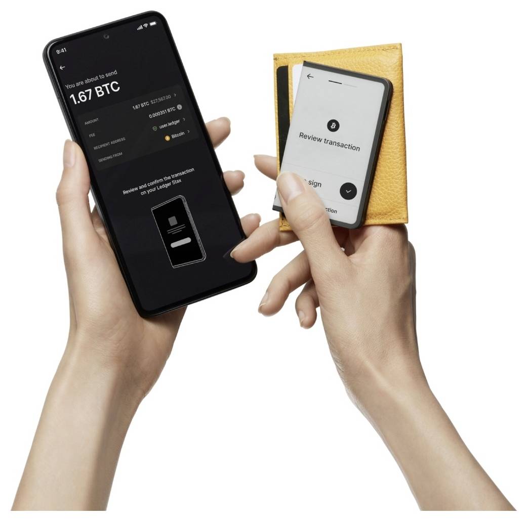 Ruka drží chytrý telefon s aplikací Bitcoin peněženky zobrazující zůstatek 1,67 BTC. Druhá ruka drží hardwarovou peněženku pro potvrzení transakce.