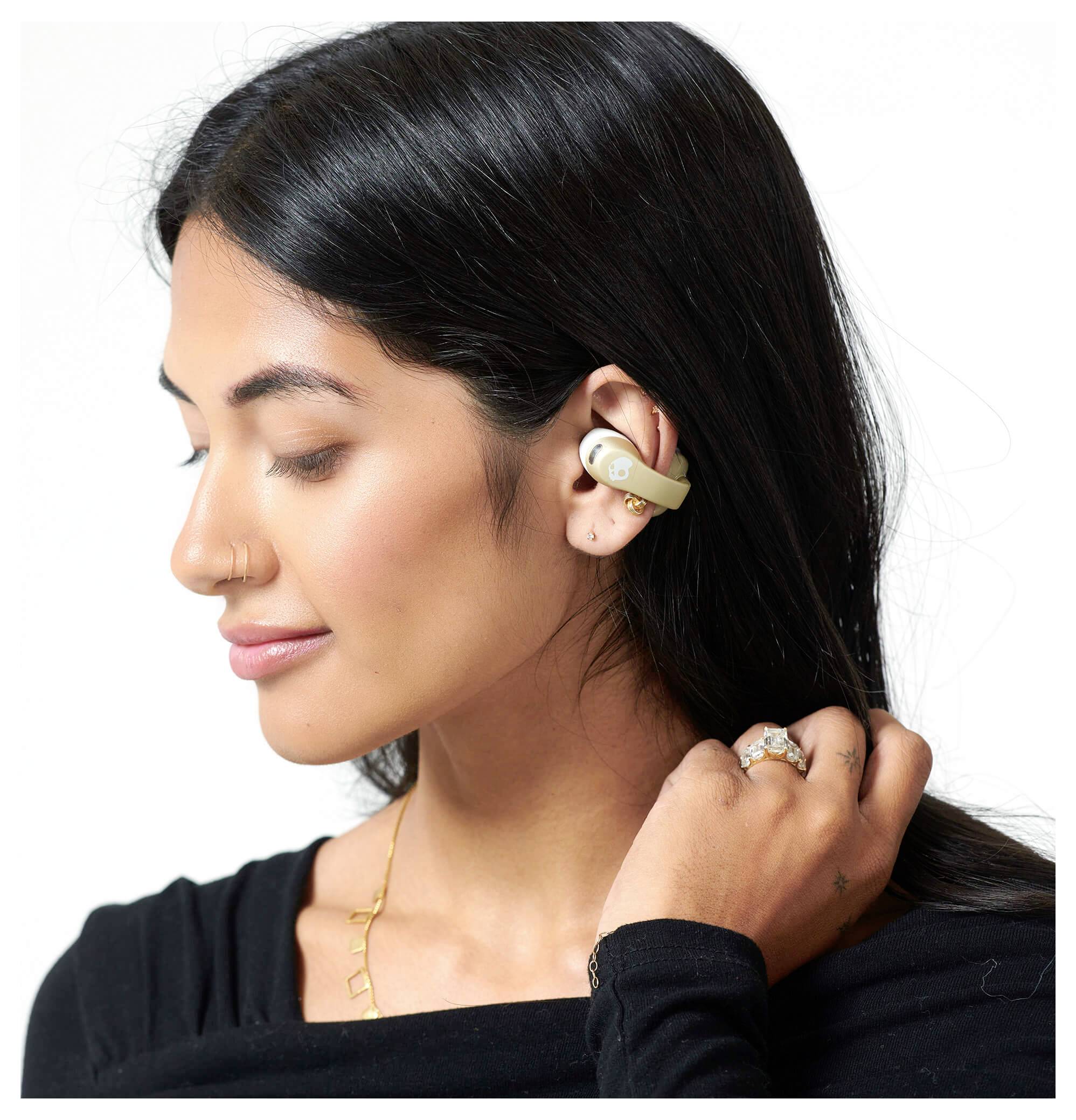 Žena s dlouhými tmavými vlasy se usmívá a nosí bezdrátová sluchátka. Je oblečena v černém topu a nosí více prstenů.