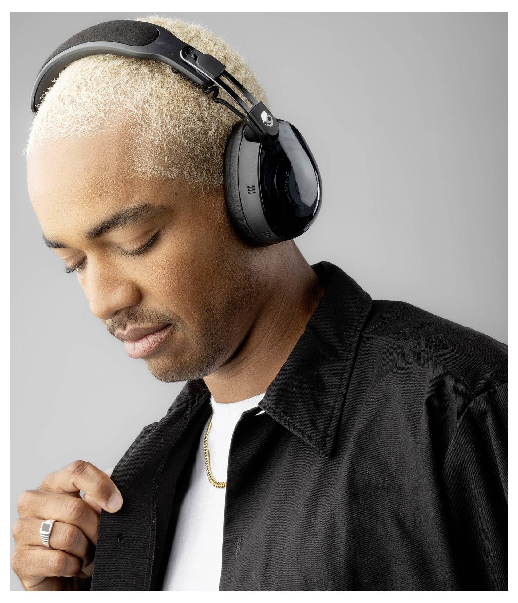 Osoba v nadrozměrných sluchátkách, pohlížející zamyšleně dolů. Jednotlivec je oblečen v černé bundě a bílé košili na neutrálním pozadí.