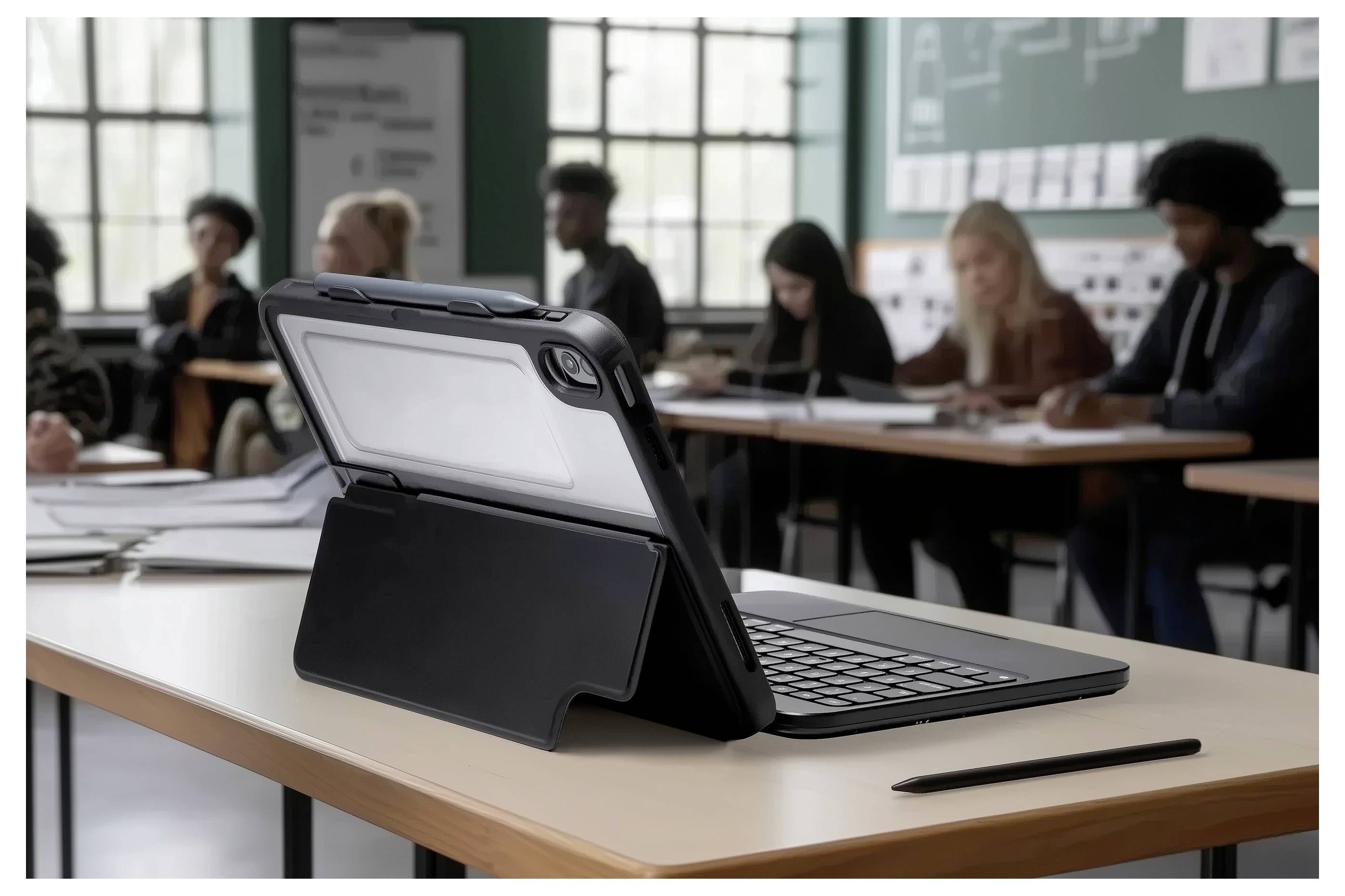 Digitální tablet s klávesnicí si leží na stole ve třídě. V pozadí sedí studenti a zabývají se studijními aktivitami.