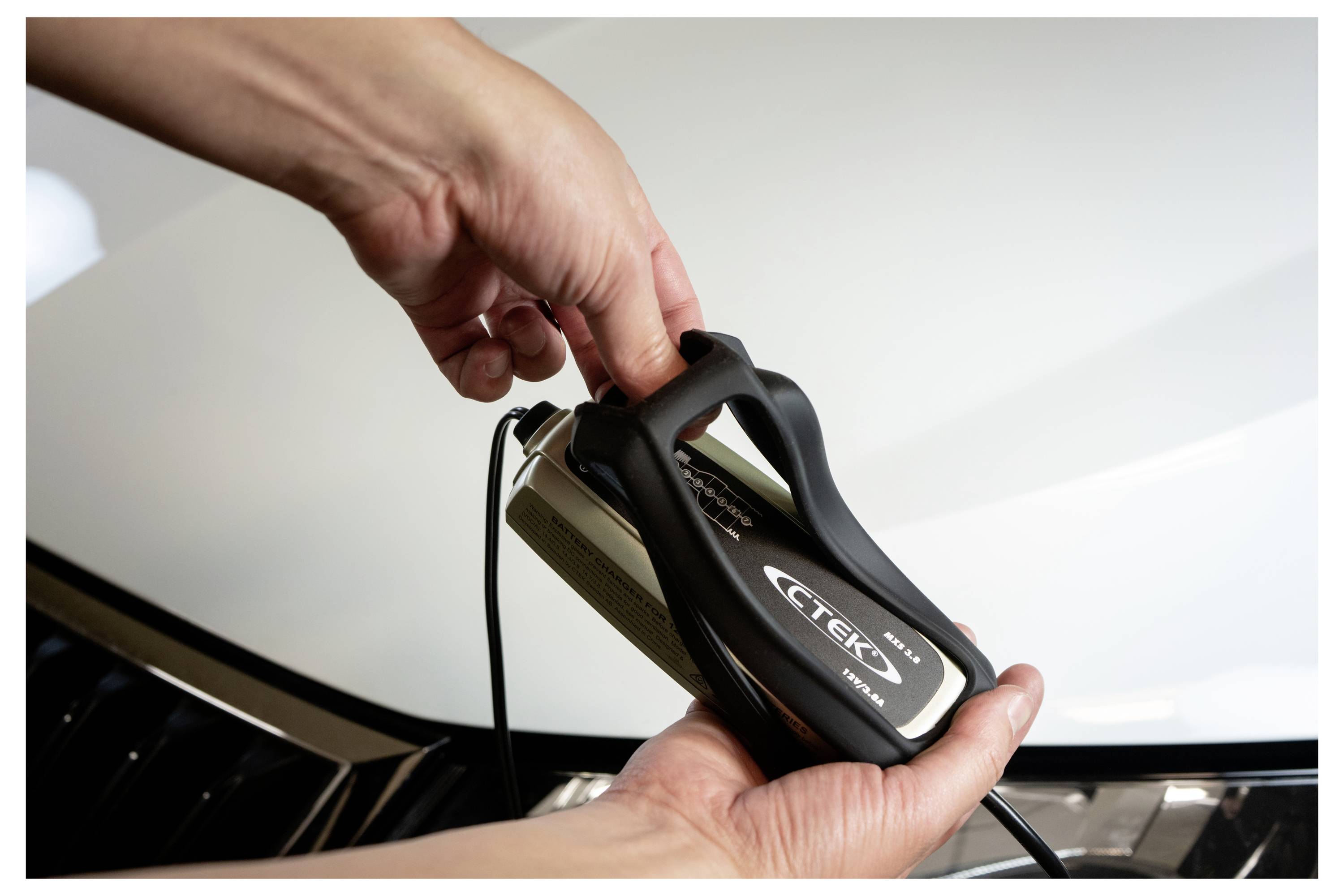 Osoba držící nabíječku automobilové baterie s logem 'CTEK', kterou nastavuje, pravděpodobně se připravuje k nabití baterie vozidla.