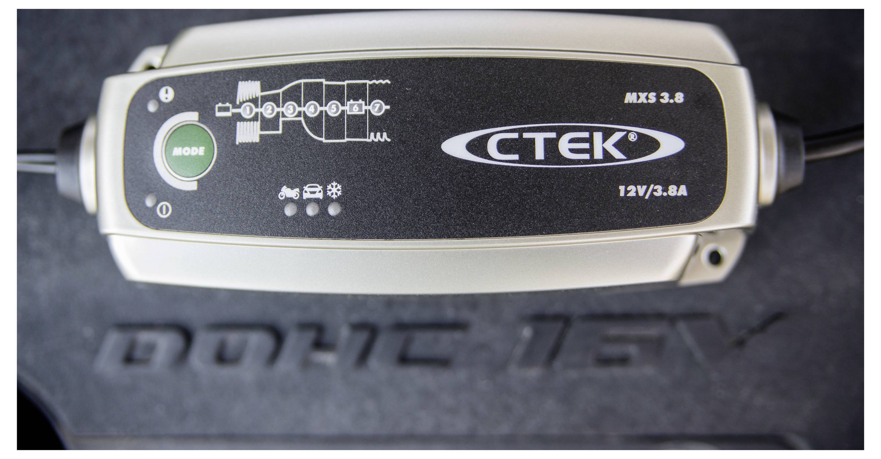 Nabíječka autobaterie označená "CTEK MXS 3.8" s tlačítkem režimu a indikátory fází nabíjení stojí na strukturovaném povrchu.