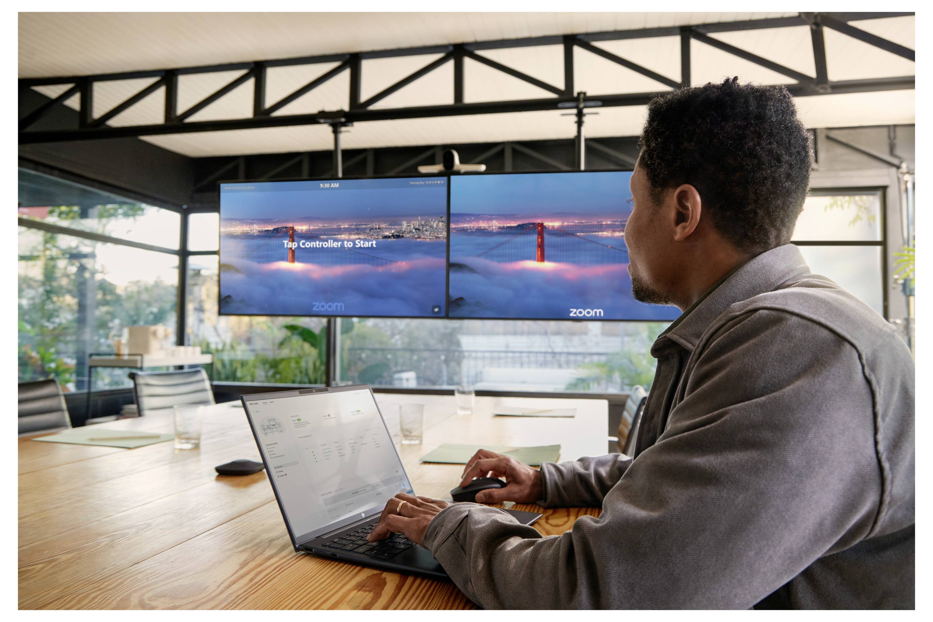 Muž používající laptop u konferenční tabulky při sledování schůzky přes Zoom na duálních monitorech zobrazujících Golden Gate Bridge.