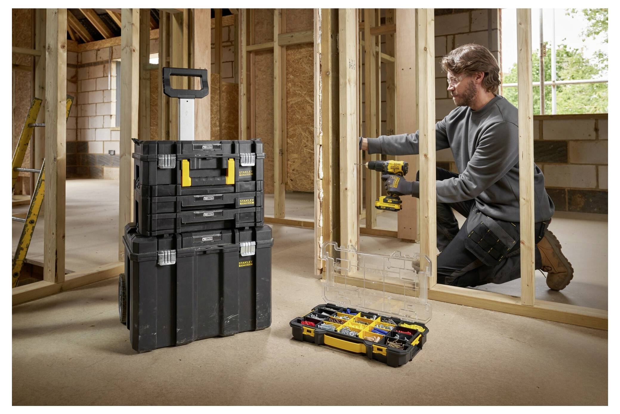 Osoba používající vrtačku konstruuje dřevěný rám v částečně postaveném pokoji. Nedaleko se nachází hromada černých a žlutých nástrojových schránek.