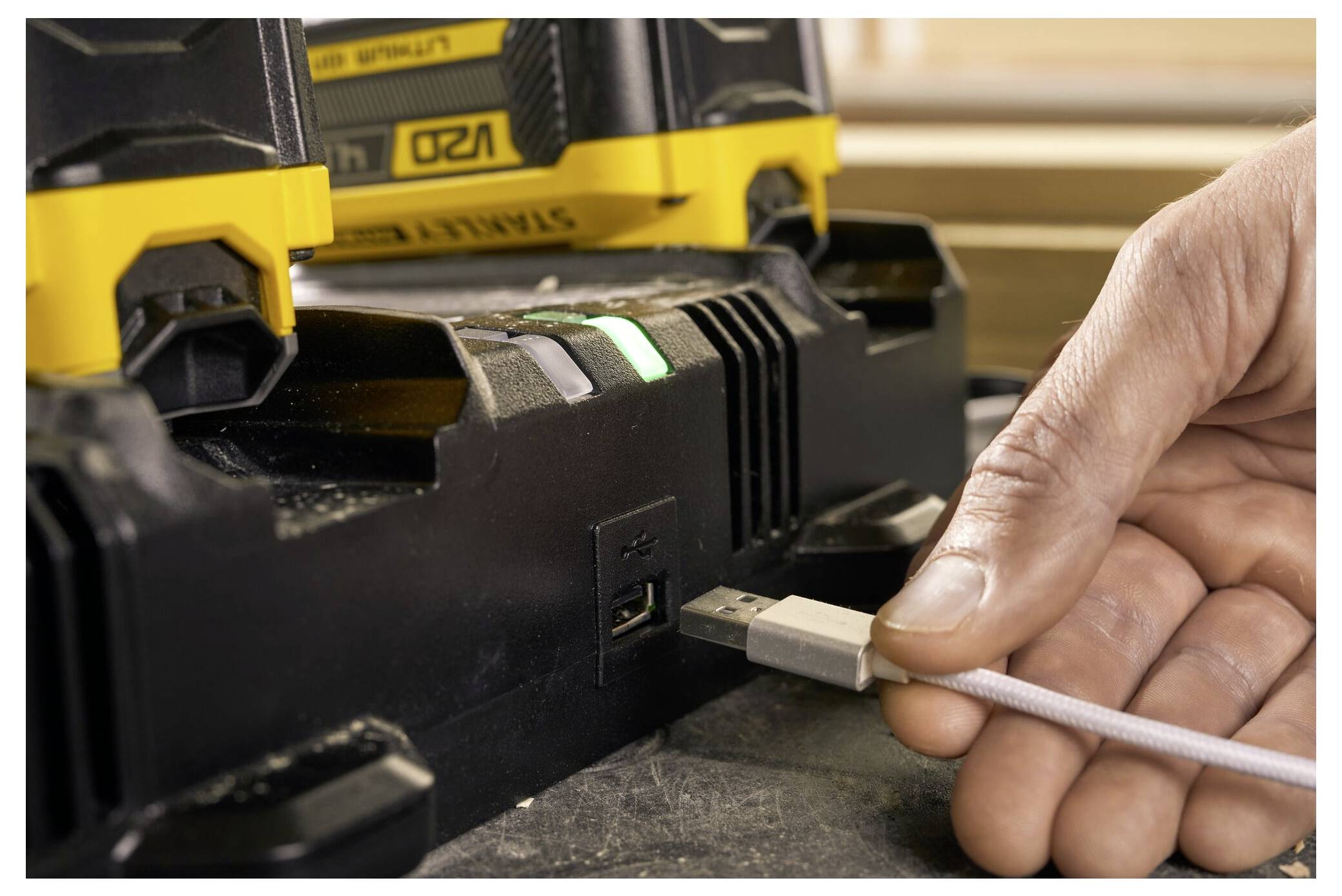 Ruka zapojuje kabel USB do nabíječky baterie elektrického nářadí, s detailním záběrem na port USB a nabíječkou zobrazující zelené světlo.