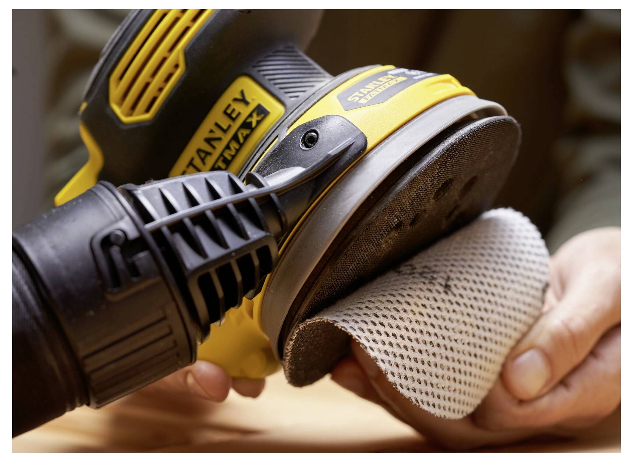 Osoba používá žluto-černou elektrickou brusku na vyhladění kusu dřeva, přičemž pevně přitiskuje nástroj na brusný papír.