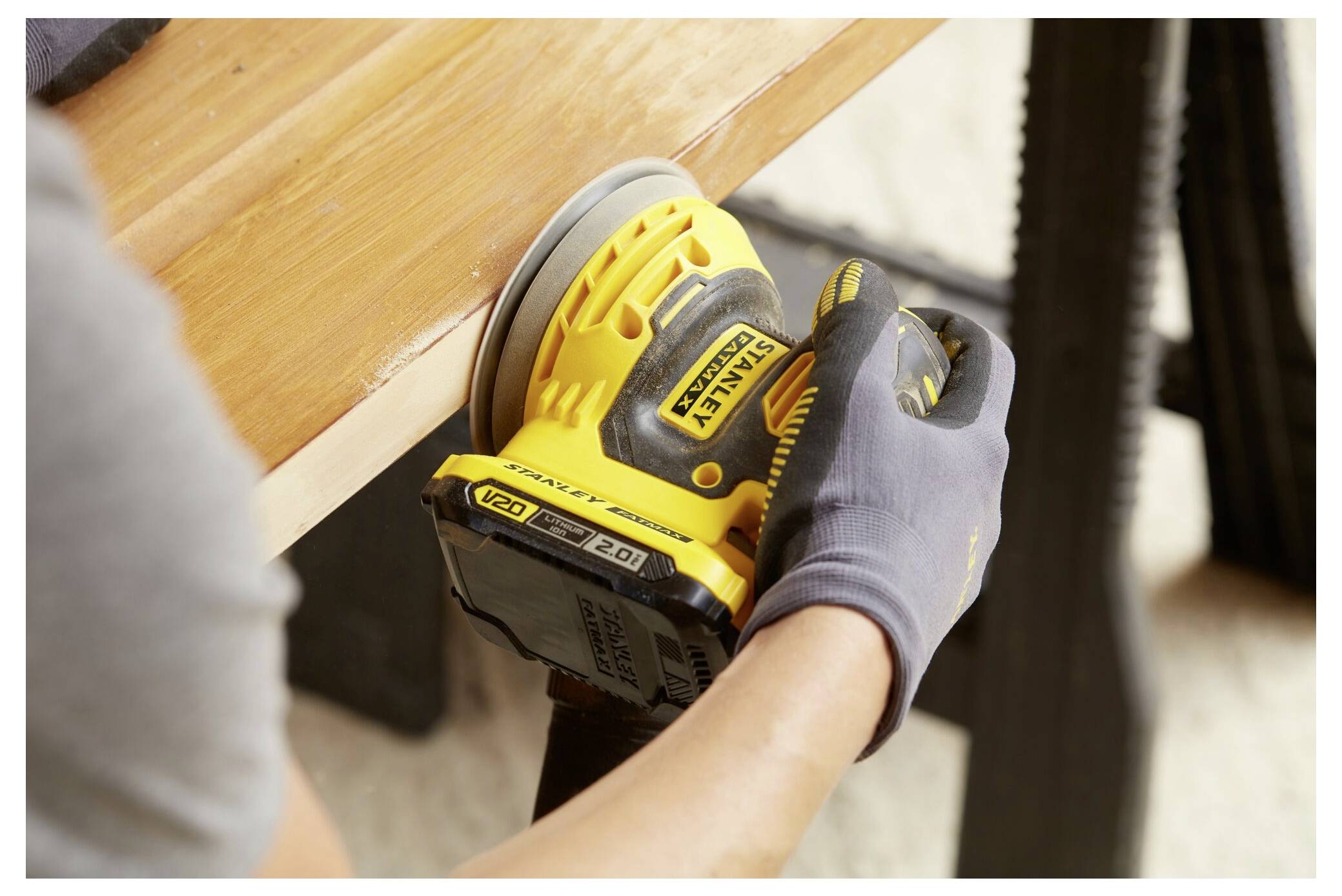 Osoba používající žlutou elektrickou brusku k vyhlazení okraje dřevěné plochy, s zaměřením na nástroj a jejich ruku v rukavici.