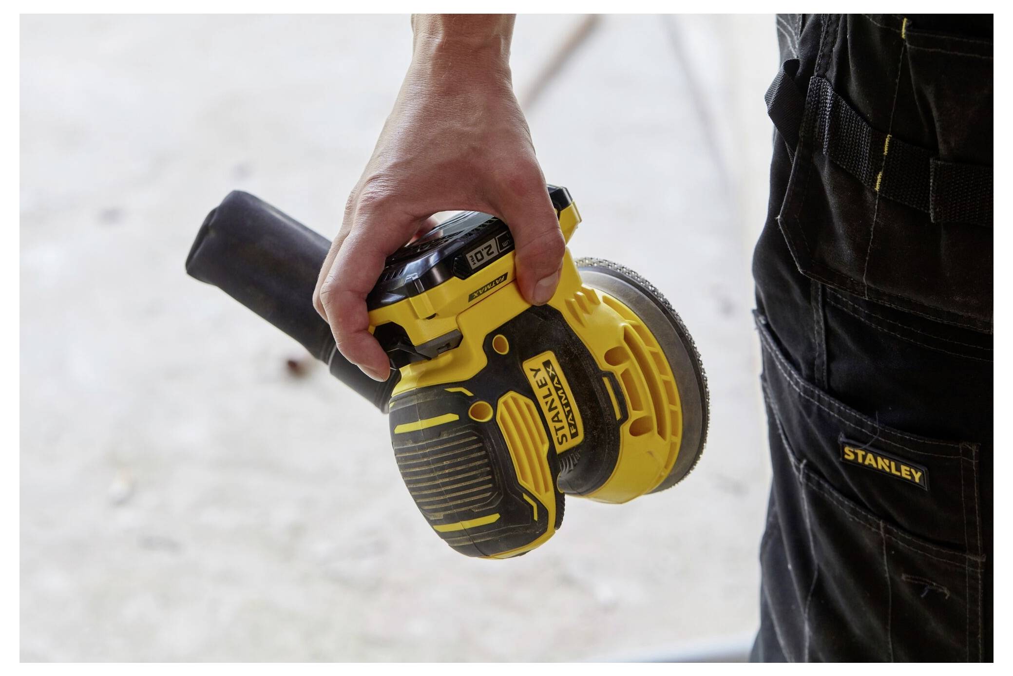 Osoba drží žluto-černý Stanley elektrickací brusku, stojící v dílně v tmavých pracovních kalhotách.