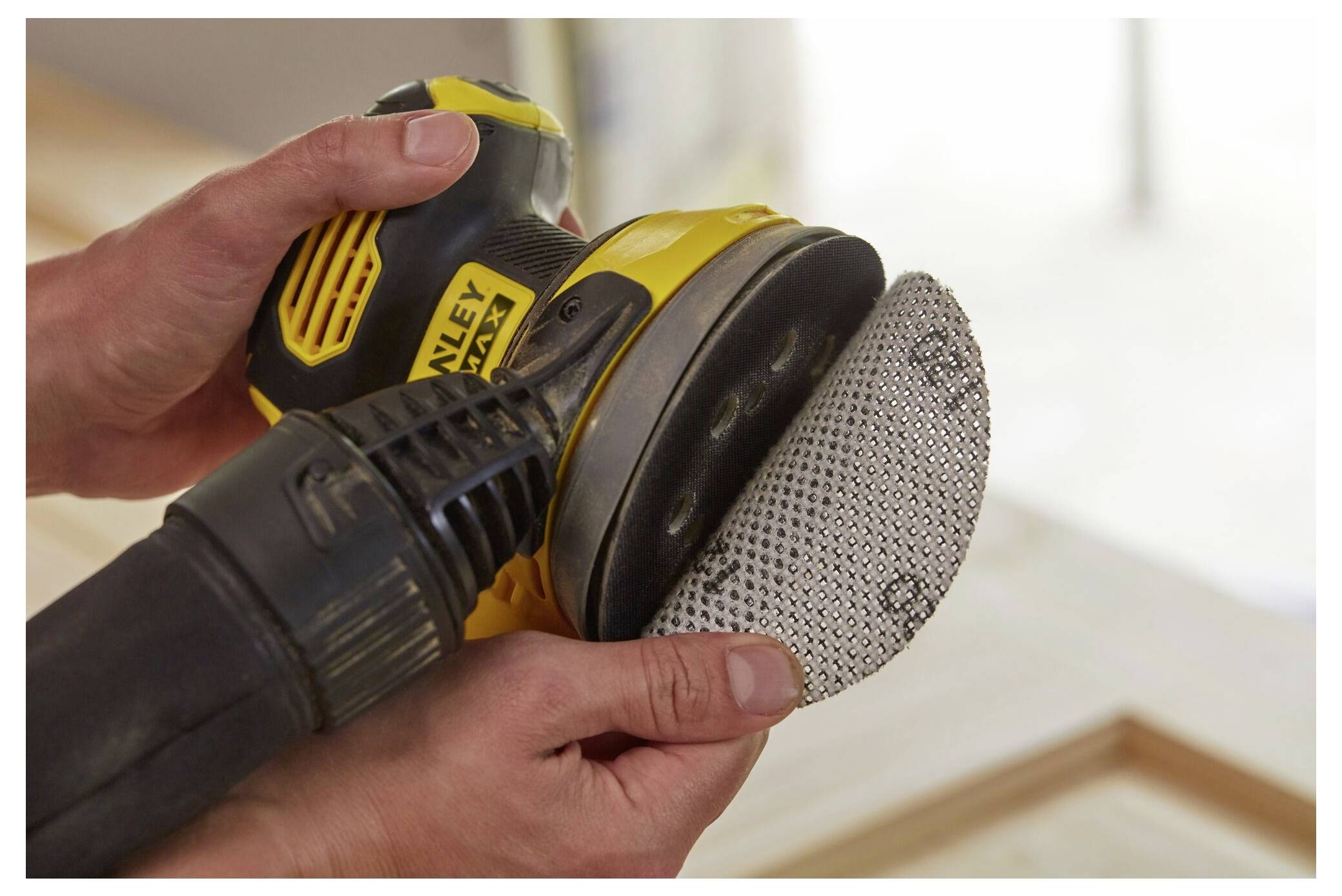 Ruce držící žluto-černou elektrickou brusku s kruhovým brusným podložem, který se právě připevňuje, což naznačuje přípravu na truhlářské práce.