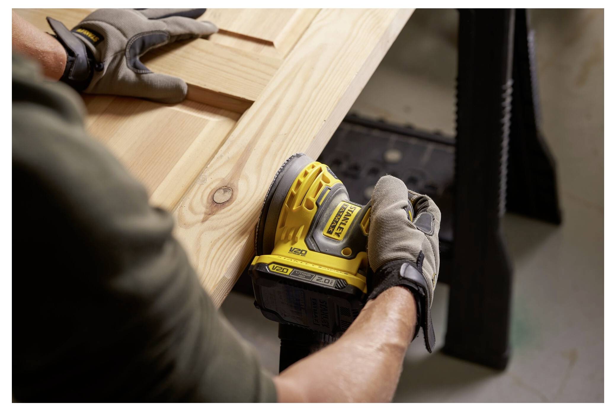 Osoba používající žlutou elektrickou brusku na vyhladění dřevěné desky na stole, nosící rukavice pro ochranu.