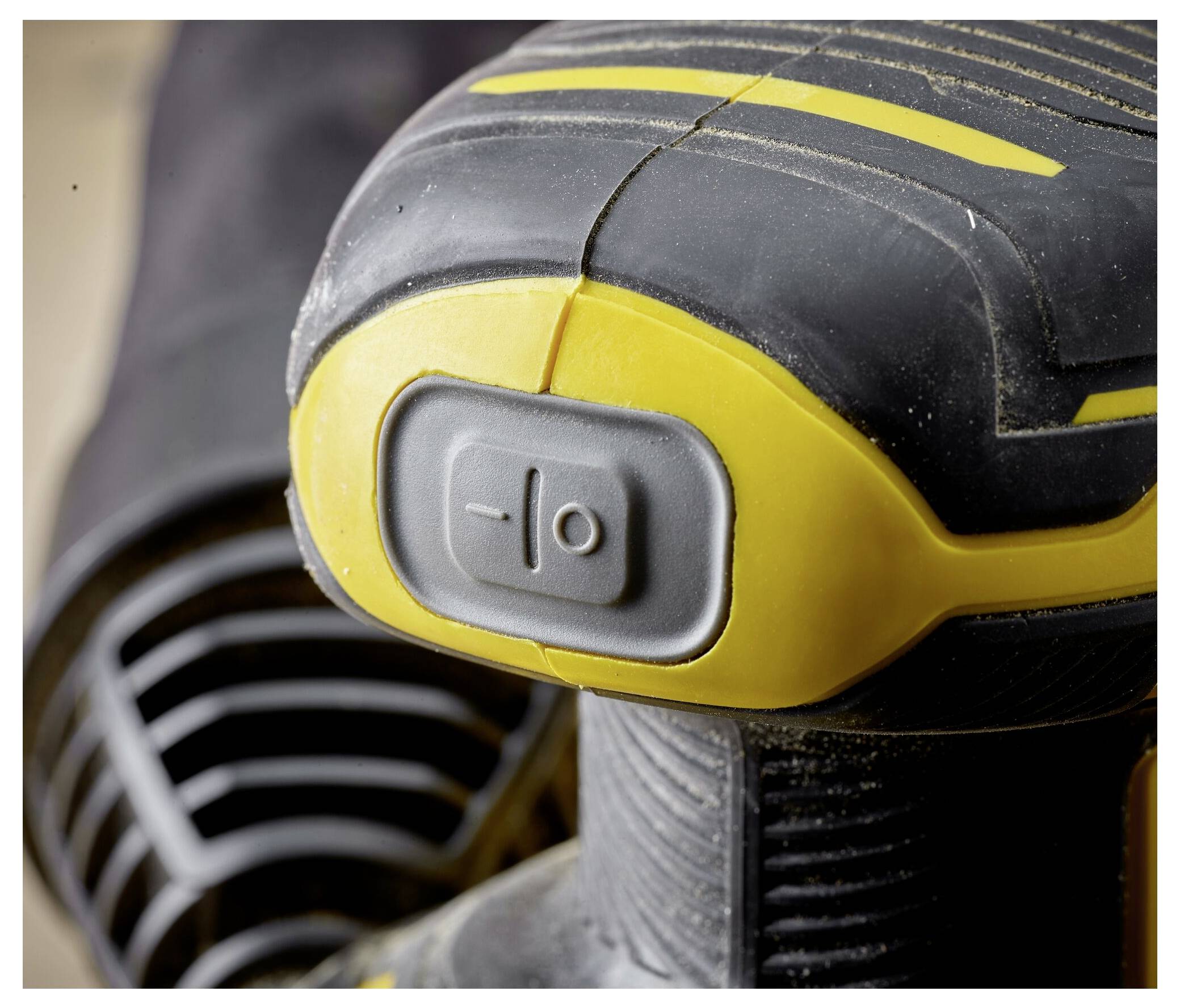 Detailní pohled na rukojeť elektrického nářadí a spínač, zobrazující strukturovanou úchopovou plochu a šedé tlačítko napájení, na rozostřeném pozadí.