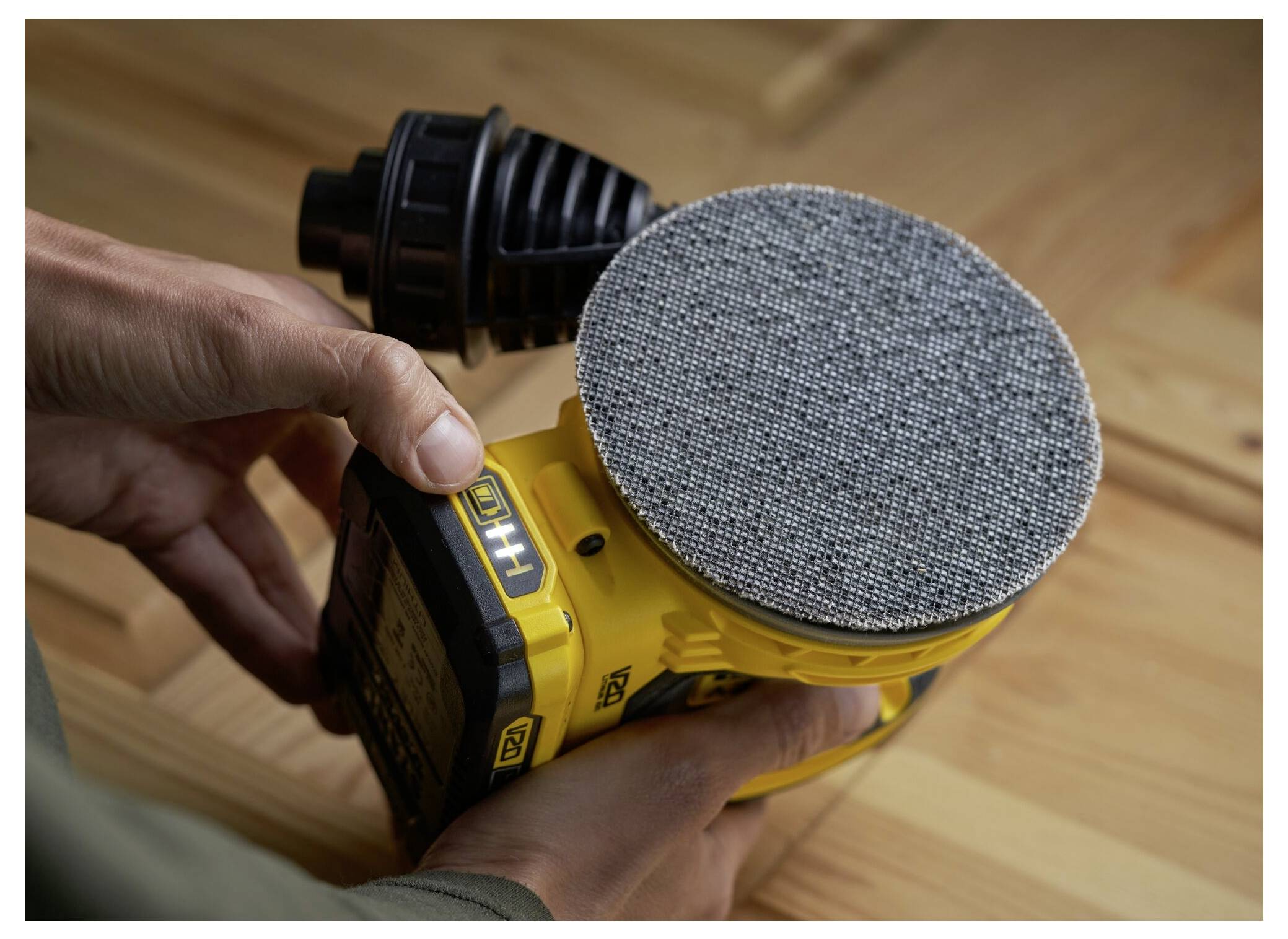 Osoba držící žlutou vibrační brusku přitisknutou na dřevěnou plochu, která ukazuje strukturovaný kruhový brusný pálek brusky.