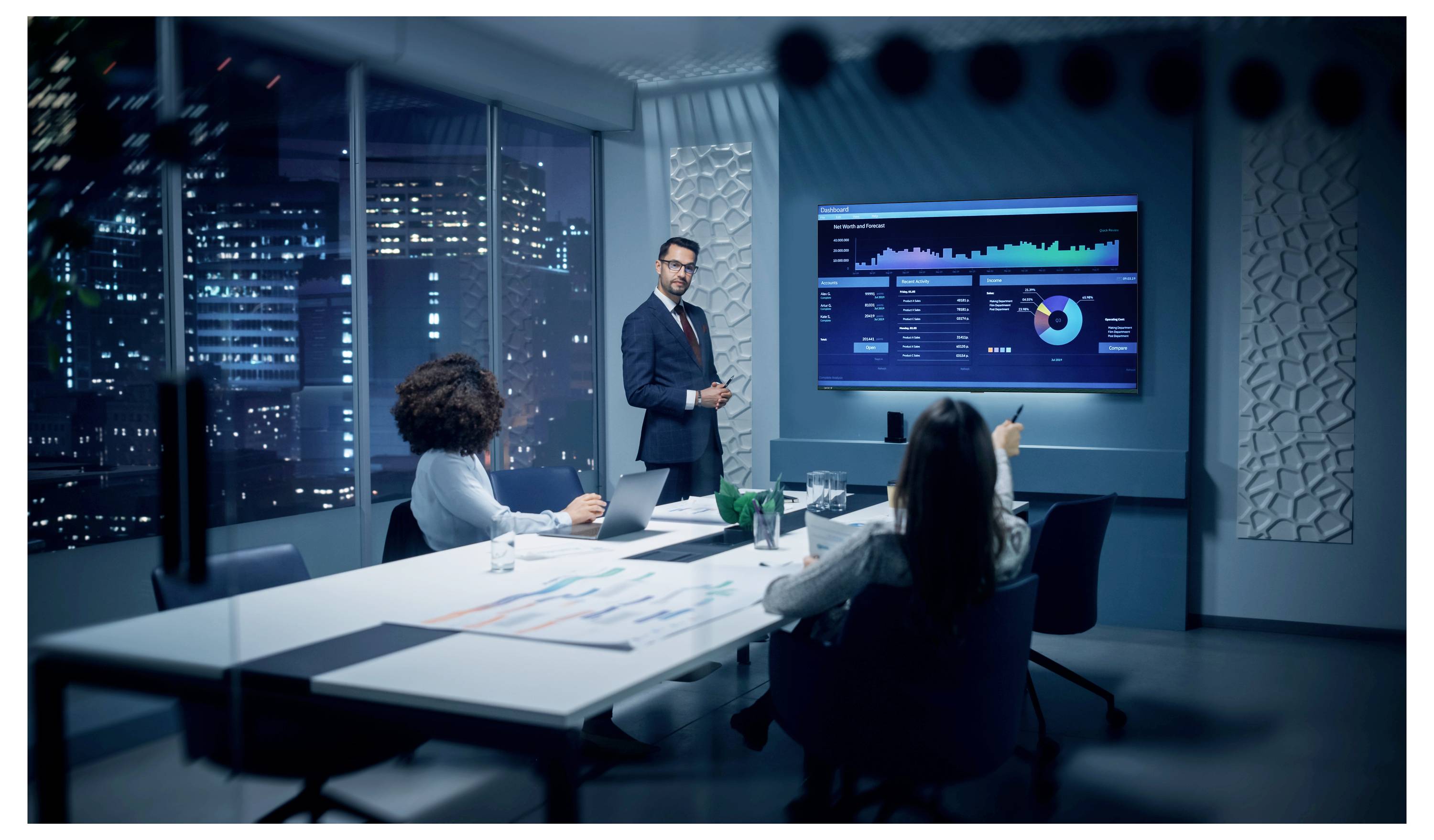 Obchodní schůze v moderní zasedací místnosti, kde prezentující ukazuje kolegům kolem stolu grafy dashboardu na nástěnné obrazovce.