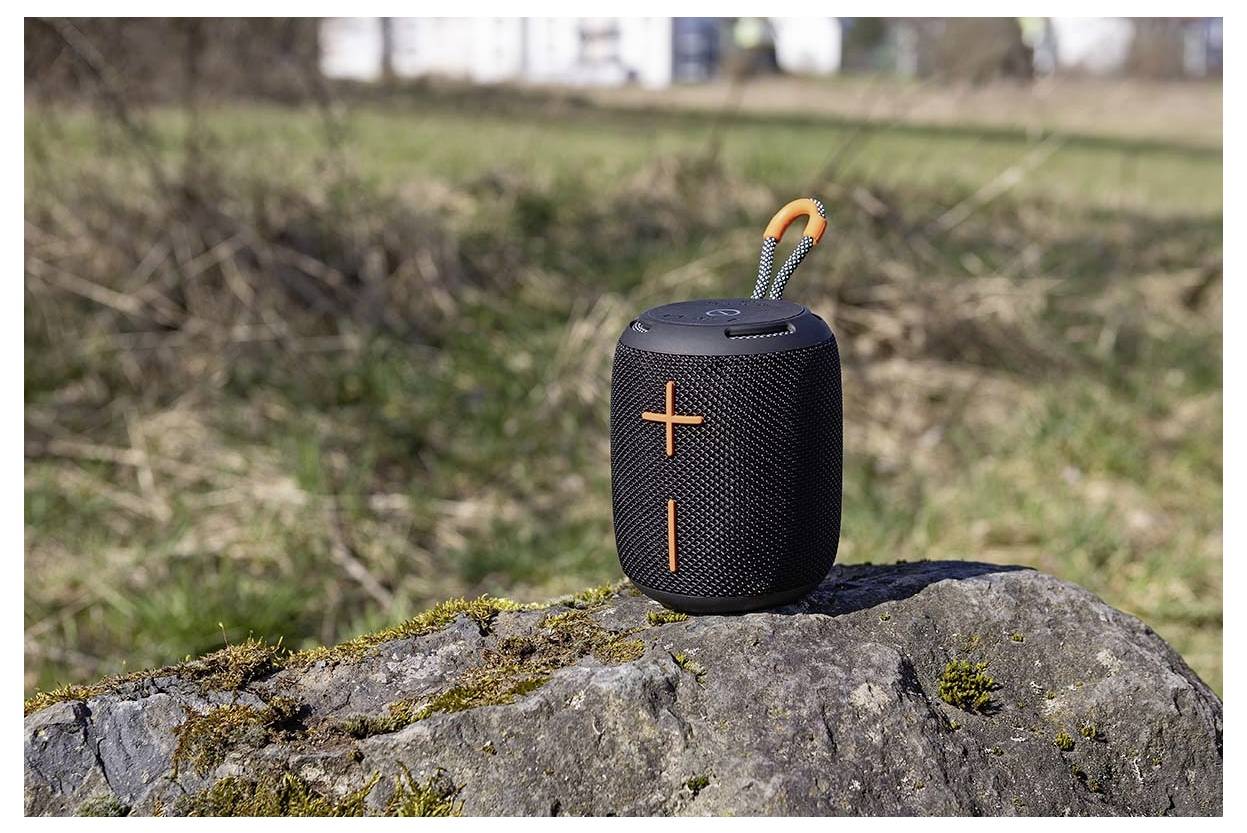 Přenosný černo-oranžový Bluetooth reproduktor sedí na mechavé skále v přírodním prostředí s rozostřenou trávou v pozadí.