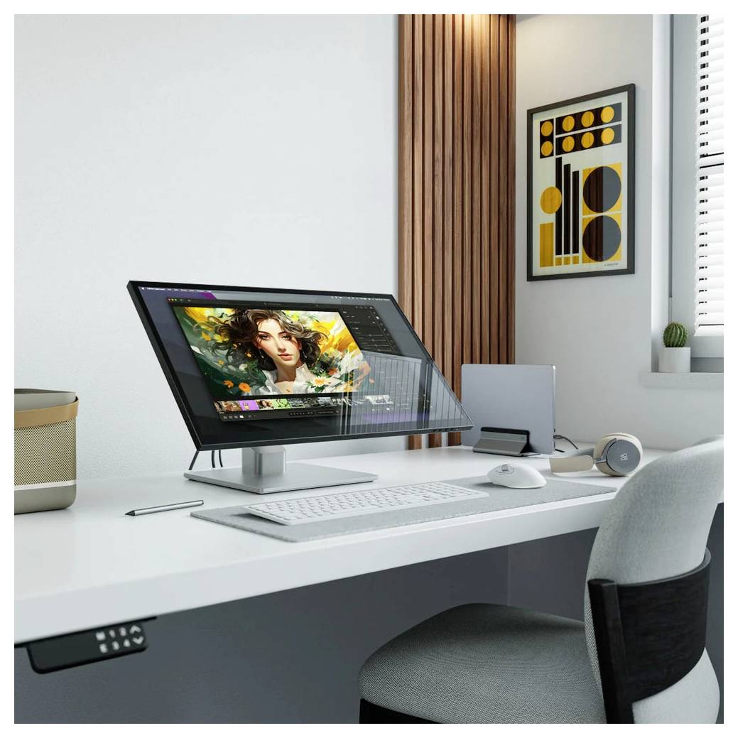 Moderní pracovní stůl s velkým monitorem zobrazujícím digitální úpravu umění, klávesnice, myš, sluchátka a minimalistické nástěnné umění v jasné místnosti.