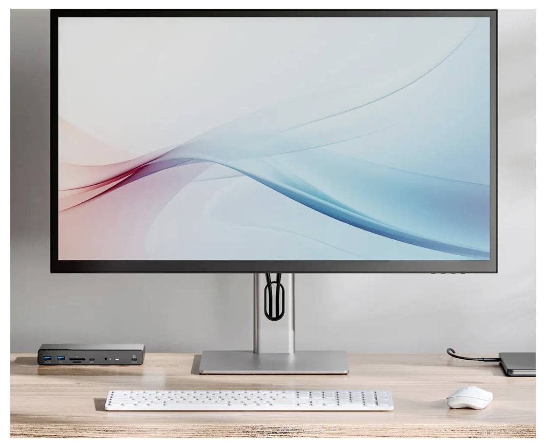 Moderní nastavení stolního počítače se širokouhlým monitorem, klávesnicí, myší a malým dokem na dřevěném stole před jednoduchou stěnou.