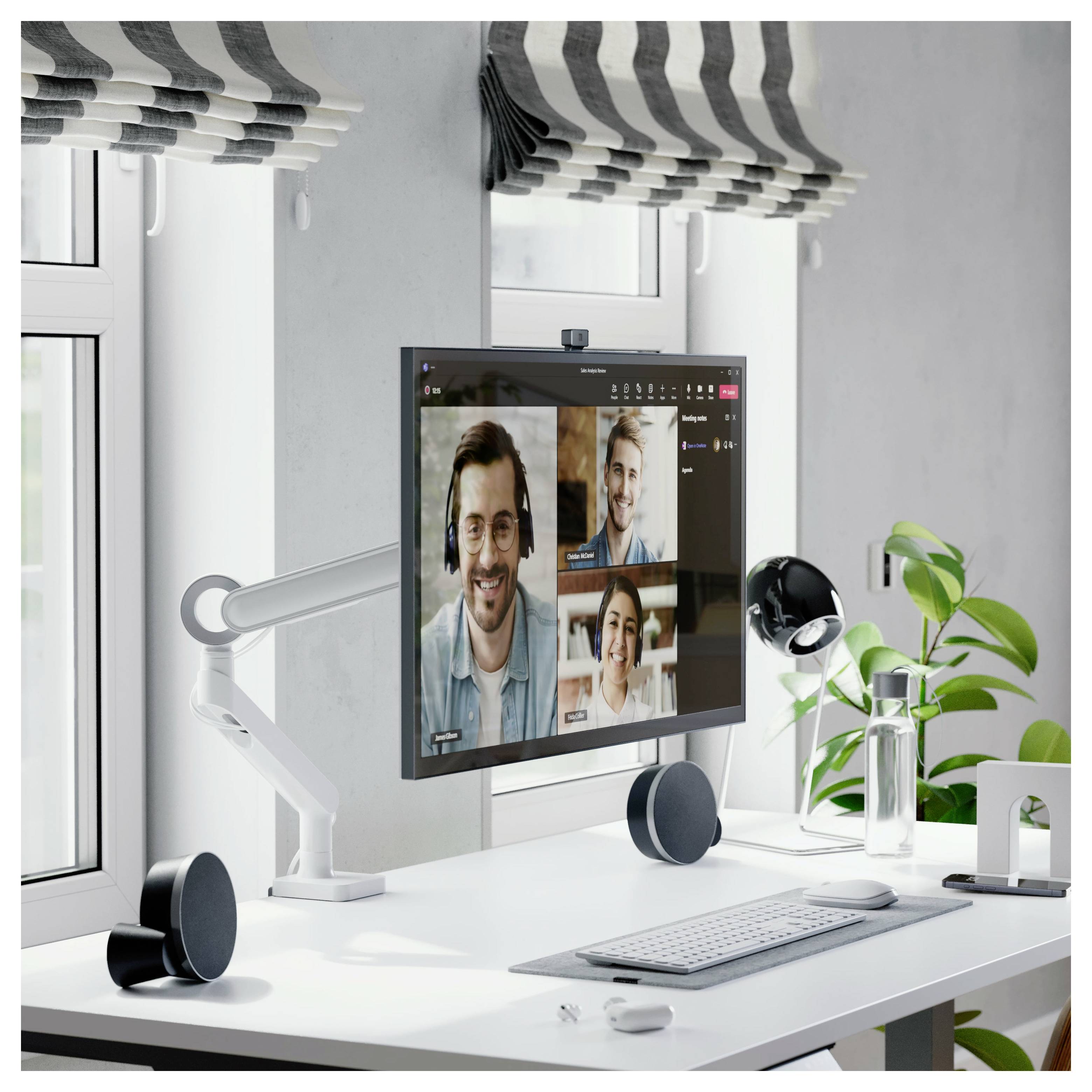 Moderní domácí kancelář s monitorem zobrazujícím videokonferenci tří osob, plus klávesnice, reproduktory, lampa a pokojová rostlina na bílém psacím stole.