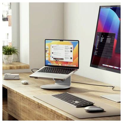 Moderní pracovní stůl se notebookem na stojanu, externím monitorem, klávesnicí a myší na dřevěné pracovní ploše u okna.