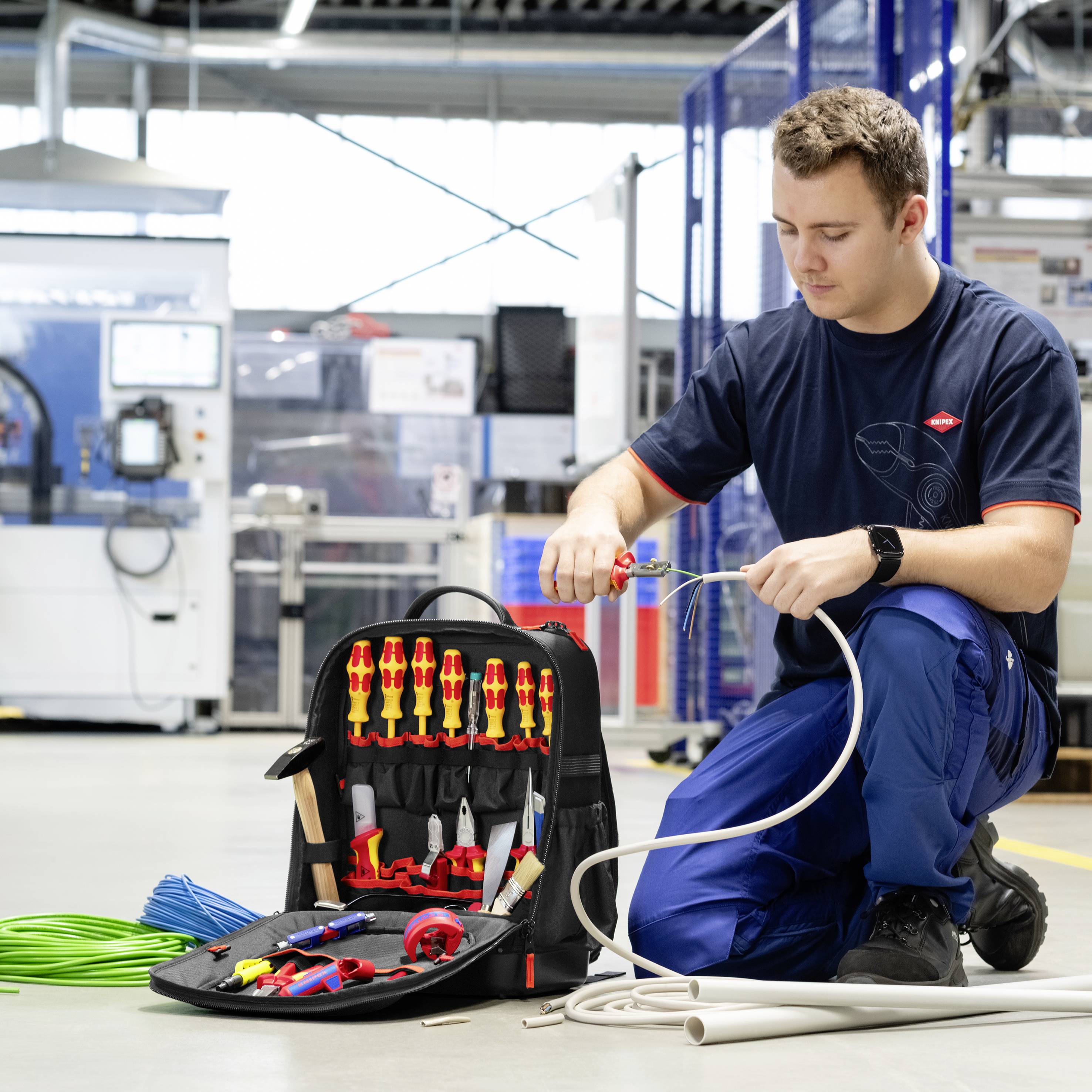 Technik klečí v dílně a testuje a zapojuje kabely vedle otevřené brašny na nářadí plné kleští, nůžek a izolovaných šroubováků.