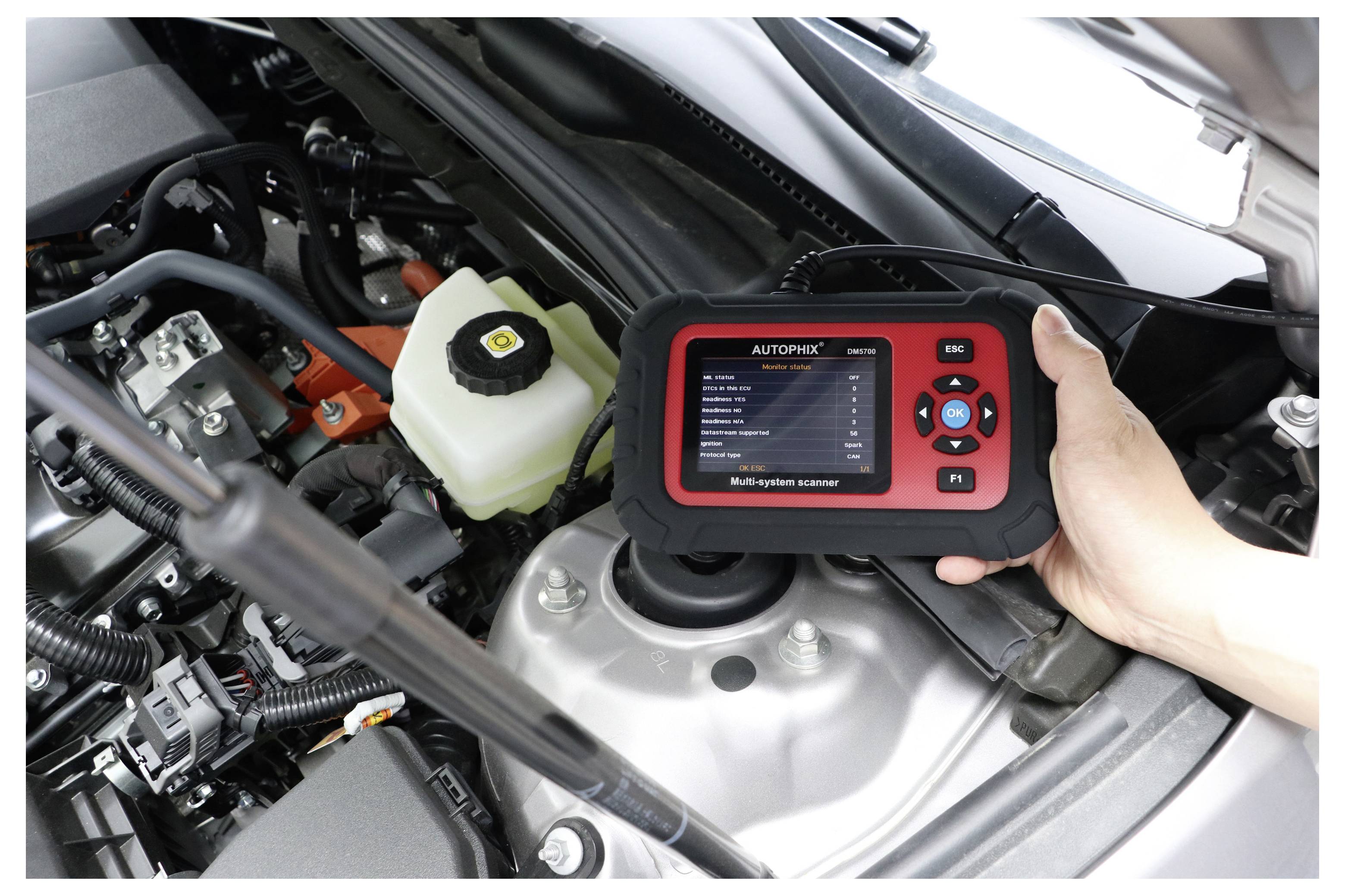 Ruka držící automobilní diagnostickou skenér Autophix nad motorem automobilu, kontrola diagnostiky poblíž baterie a nádoby brzdové kapaliny.