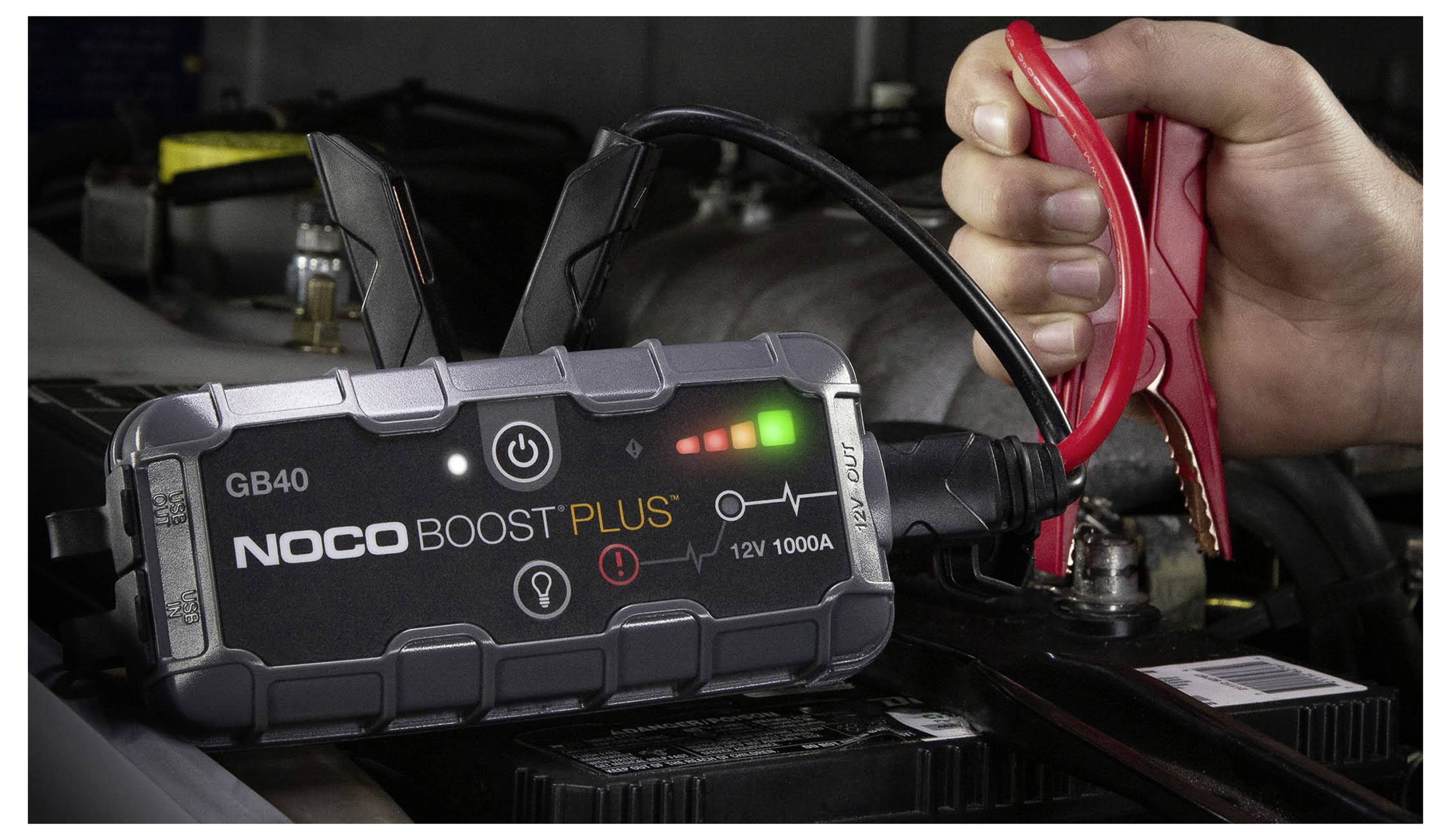 Ruka připojuje červené přísavné kabely k autobaterii pomocí přenosného startovacího zařízení NOCO Boost Plus GB40 v motorovém prostoru vozidla.