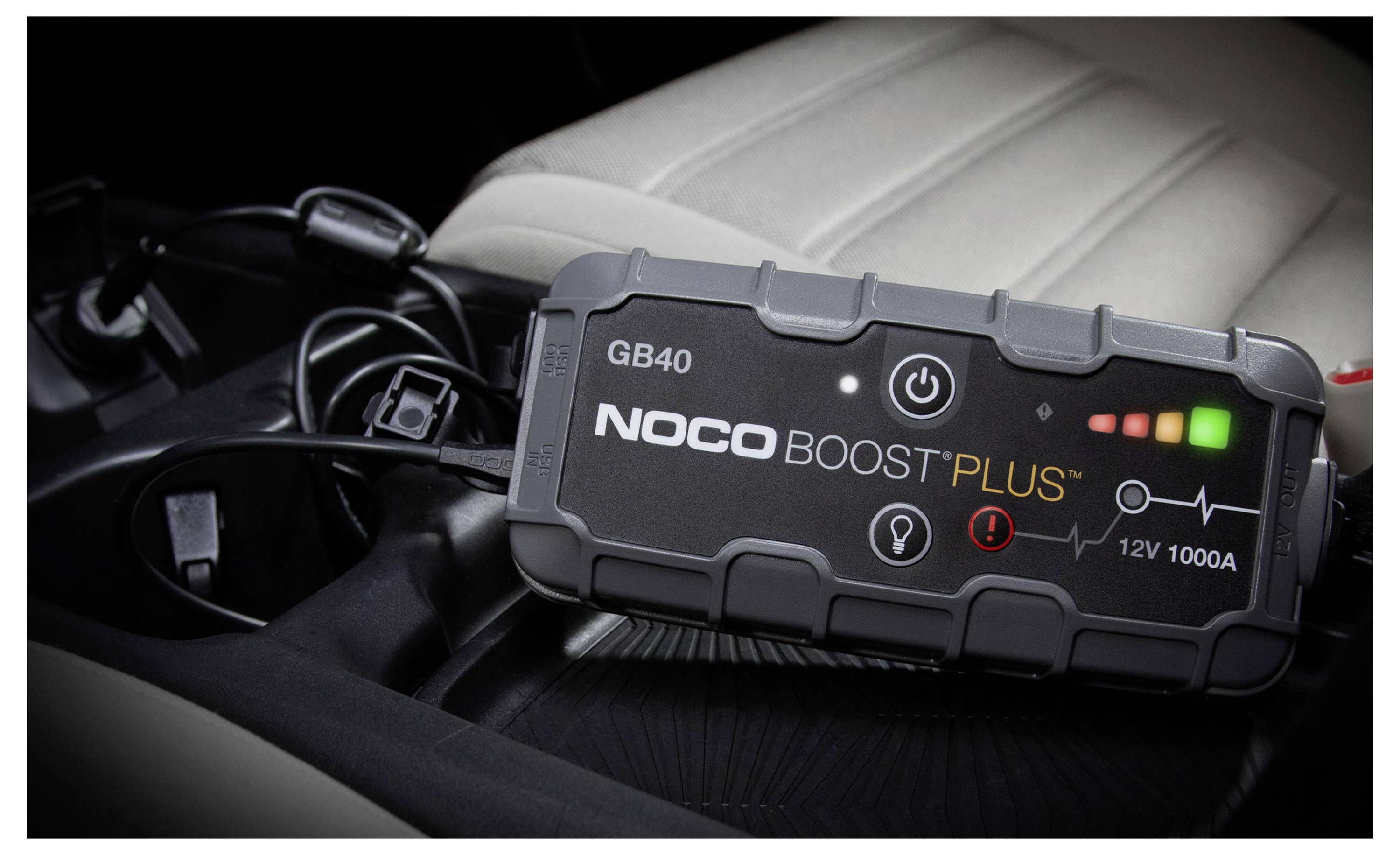 Přenosný startér NOCO Boost Plus GB40 s osvětlenými indikátory napájení leží na autosedačce s připojenými kabely poblíž.